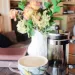 A floral coffee mug next to a french press coffee maker on a glass countertop next to a white flower vase with dried pink flowers in it next to red books for post Why French Press Coffee Does Taste Better (Recipe).