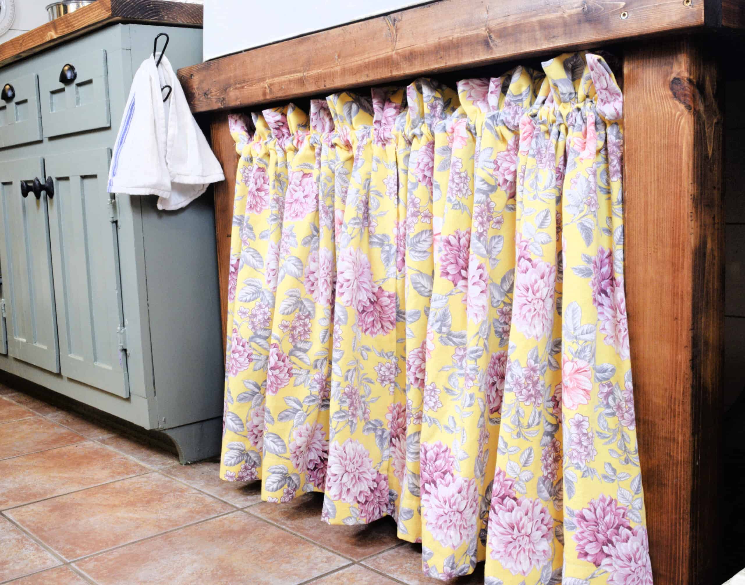 Brown sink frame with a yellow and pink floral cotton material sink skirt attached. Next to a green cabinet with a brown countertop on top of brown tile.