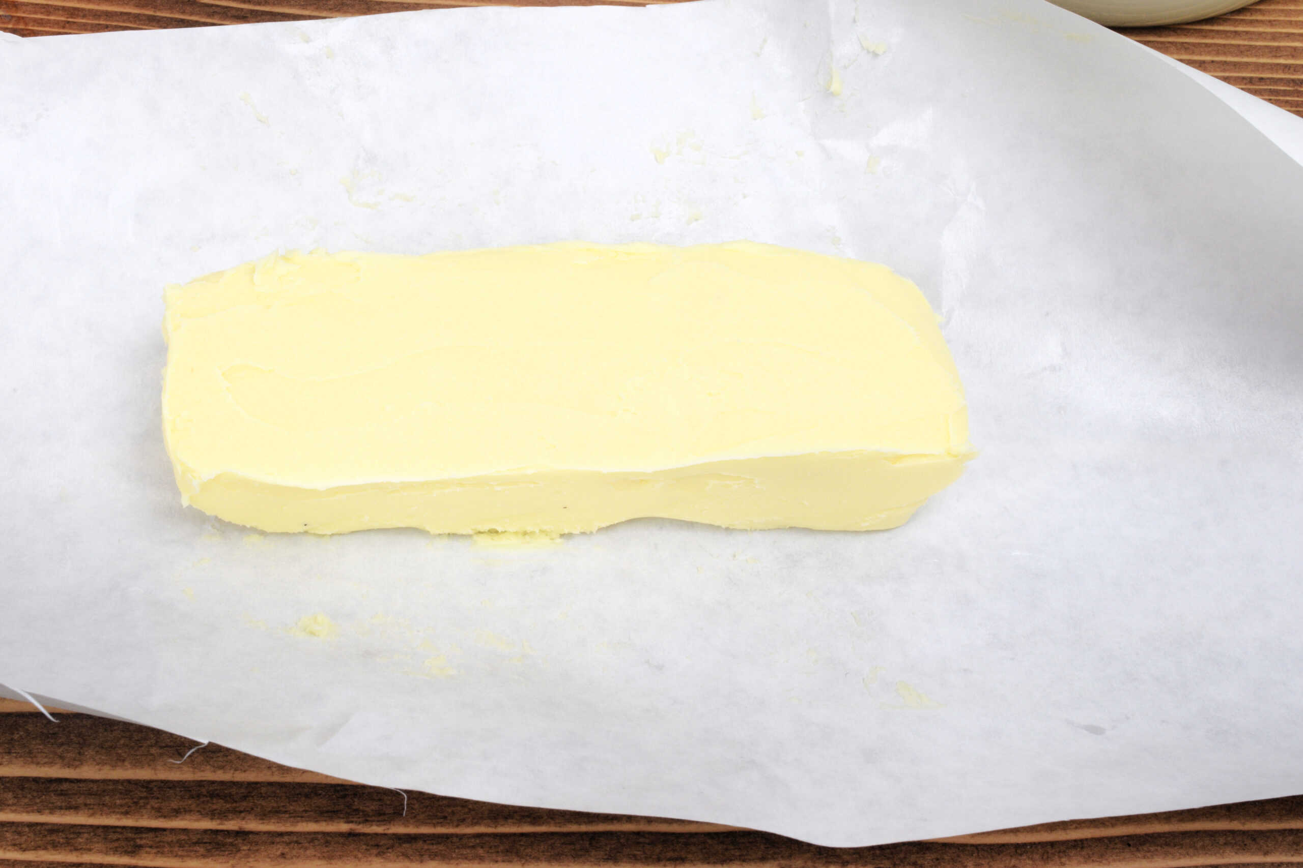 Smooth block of freshly shaped homemade butter resting on a sheet of parchment paper over a rustic wooden countertop. The pale yellow butter has clean, even edges, highlighting the final stage of making butter from raw milk in a homestead kitchen.