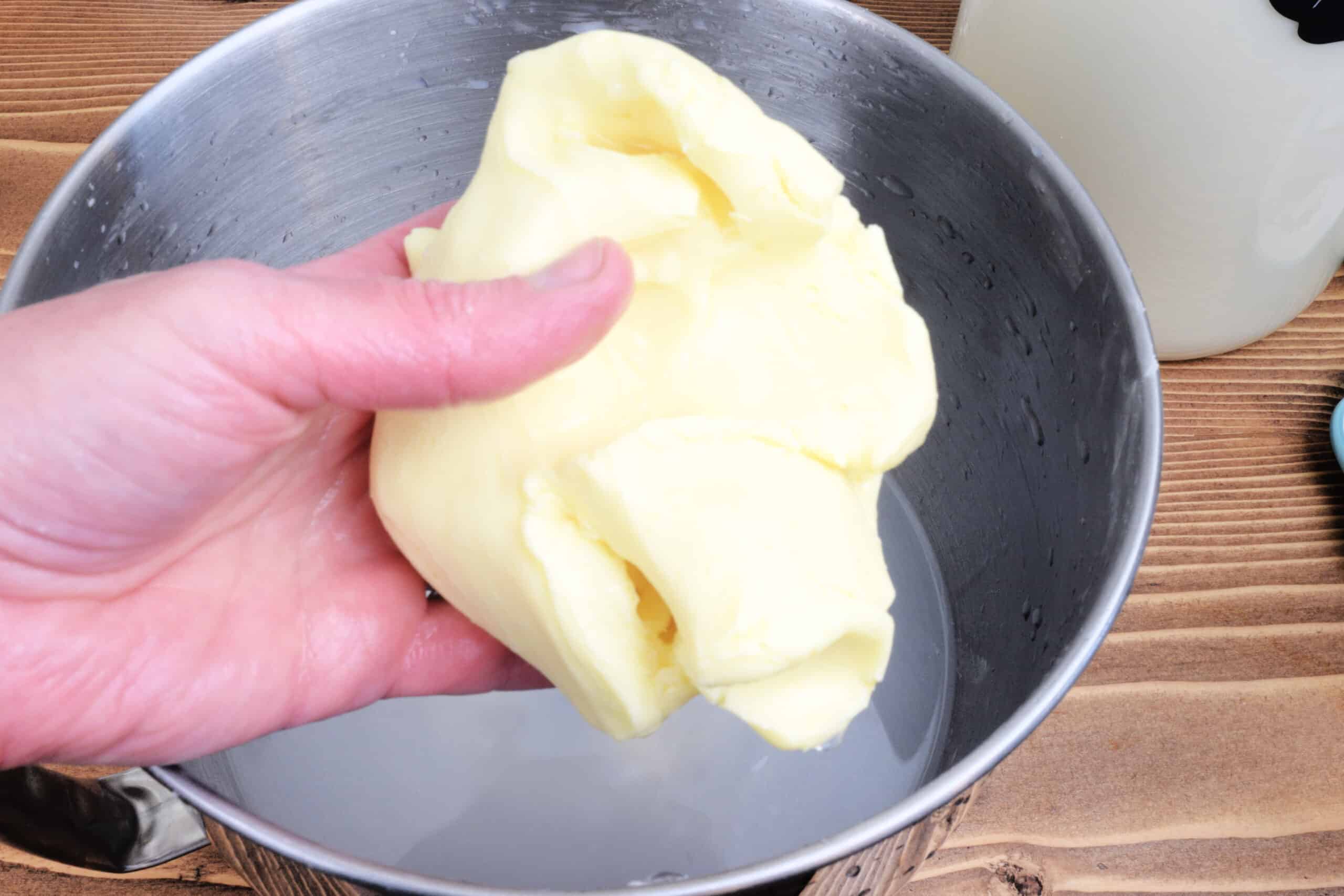 Hand holding a soft mound of freshly washed homemade butter over a stainless steel bowl filled with cold water. The bright yellow butter has a smooth, pliable texture, set in a rustic homestead kitchen environment with jars of raw cream nearby.