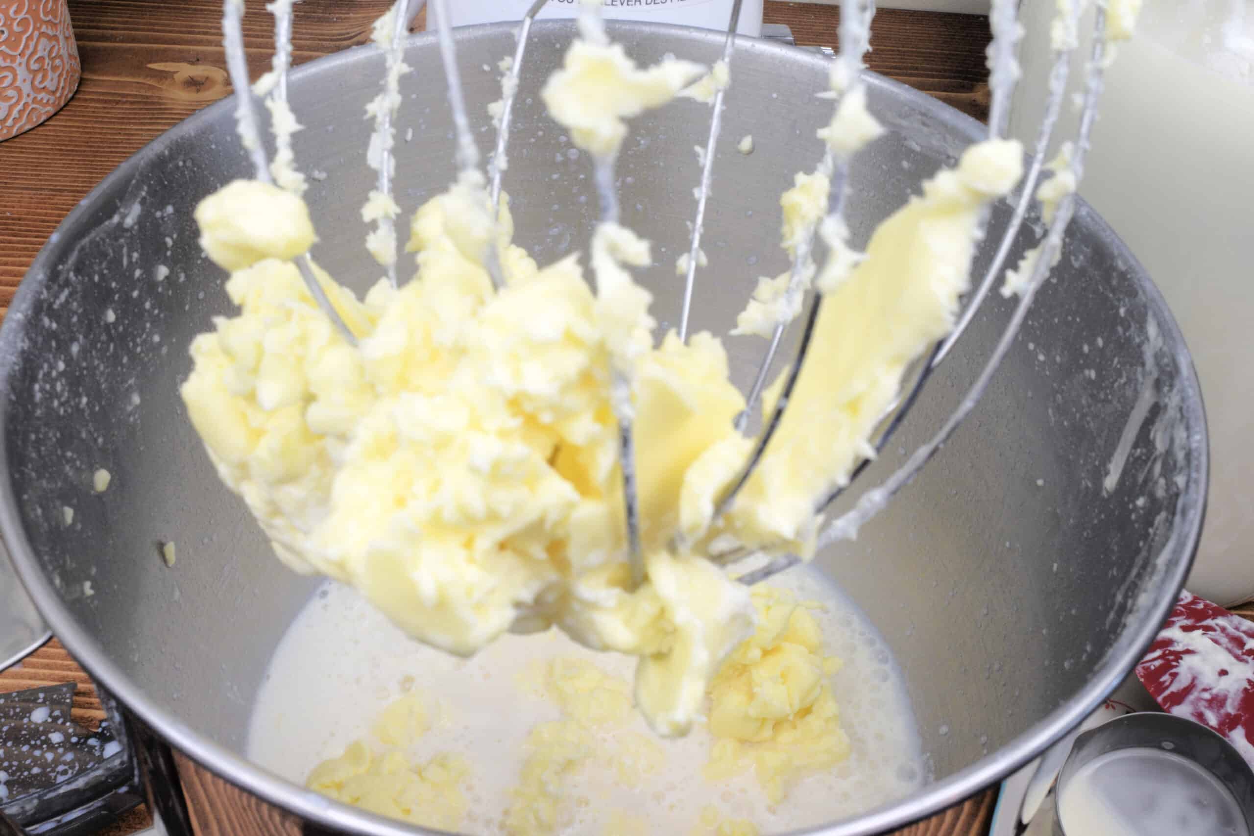 Freshly churned homemade butter clinging to the whisk attachment of a stand mixer, with creamy buttermilk pooled in the stainless steel bowl below. The bright yellow butter curds show the final stages of separating raw cream, set against a rustic wooden countertop for an old fashioned butter making tutorial.