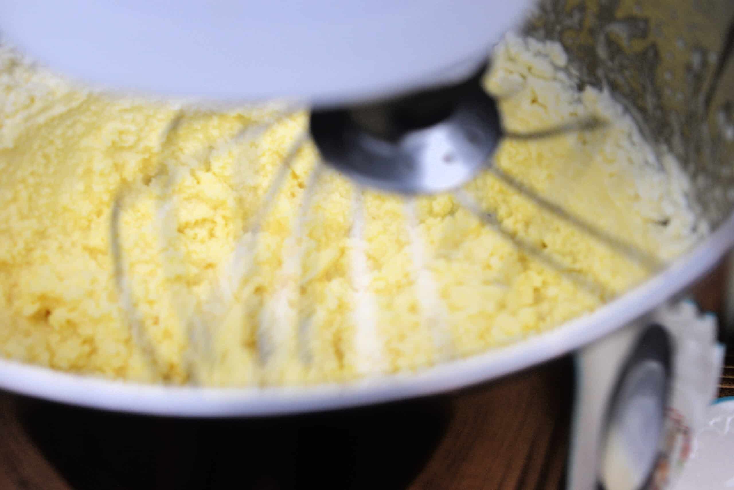 Close up of a stand mixer whisk churning raw cream into bright yellow homemade butter inside a stainless steel bowl. Soft curds are forming around the sides as the butter separates, capturing a rustic homestead kitchen moment in the butter making process.