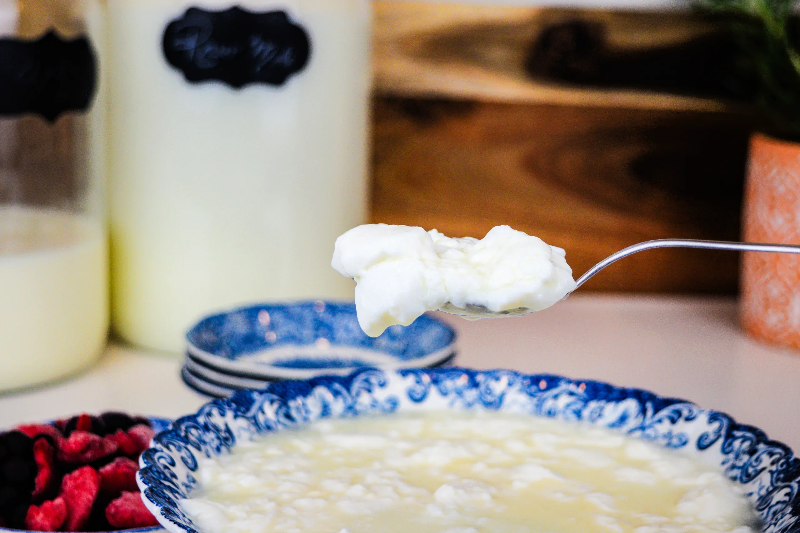 Homemade raw milk clabber on a spoon over a blue and white bowl with berries and jars of raw milk in the background.