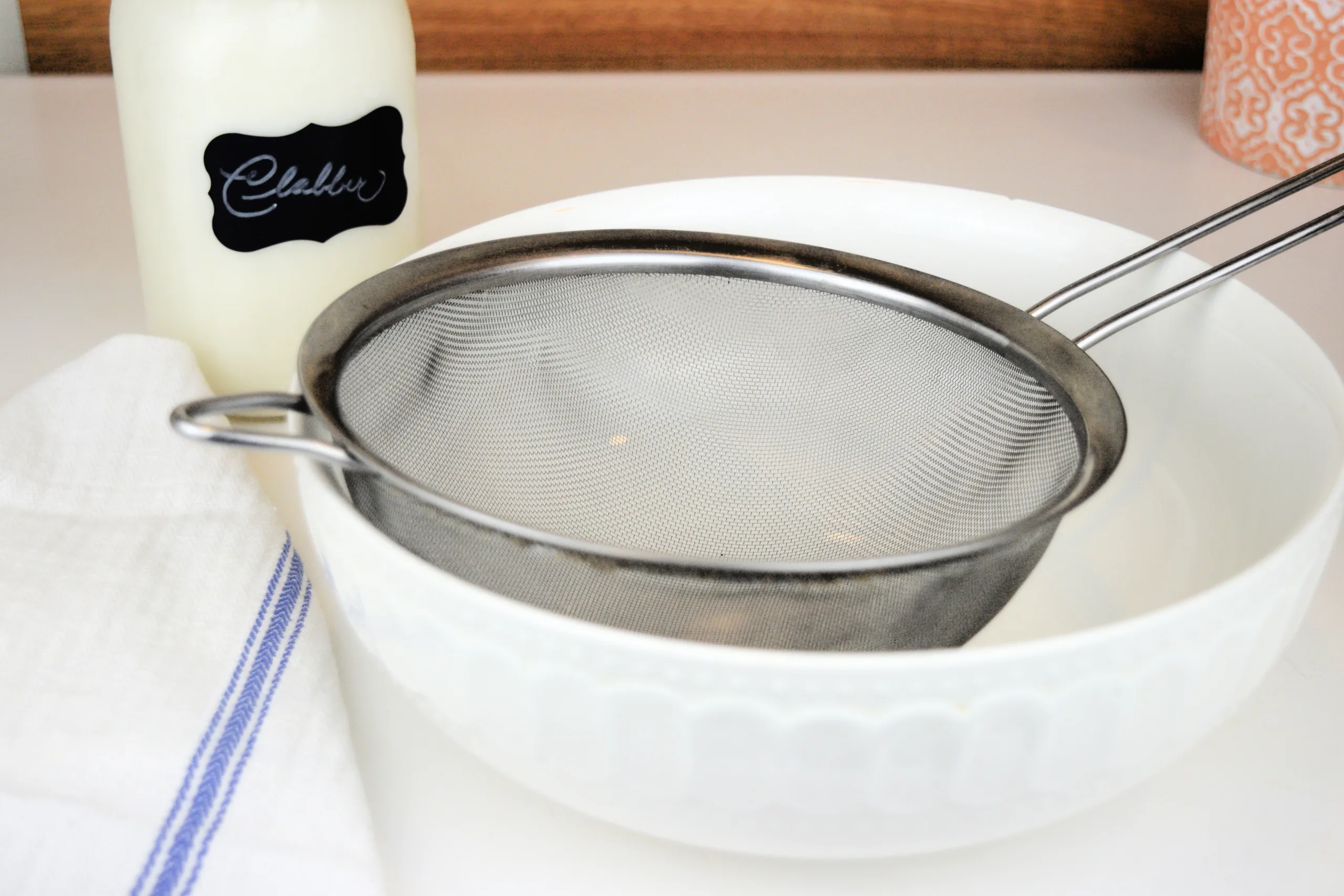 Fine mesh strainer set over a bowl with a jar of homemade clabber in the background.