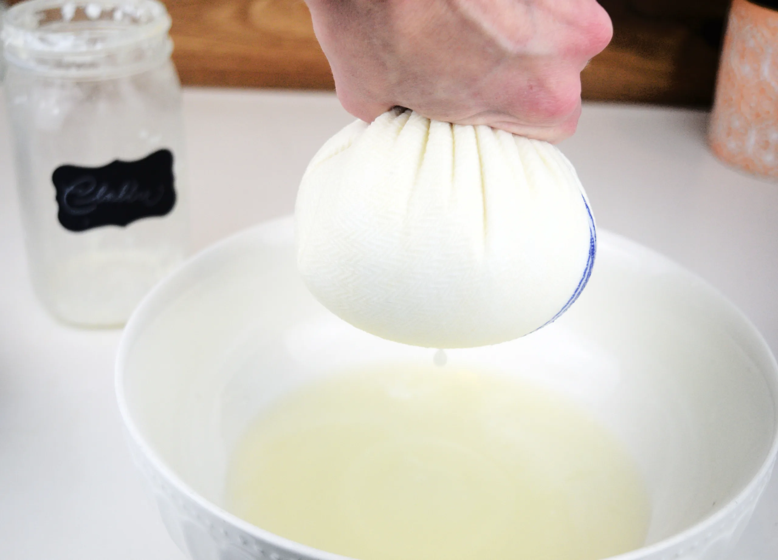 Raw milk clabber being strained through a tea towel with whey dripping into a white bowl.