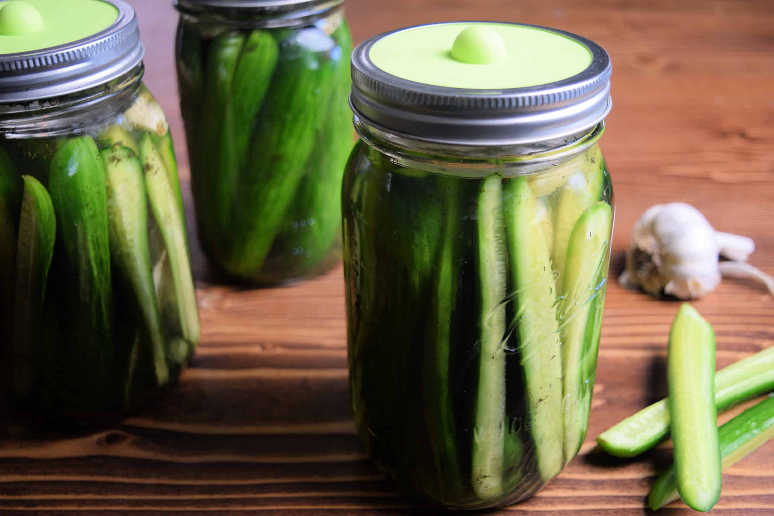 Sliced cucumbers with garlic and dill in a mason jar next to cucumbers and garlic with a fermentation weight and green burping lid.
