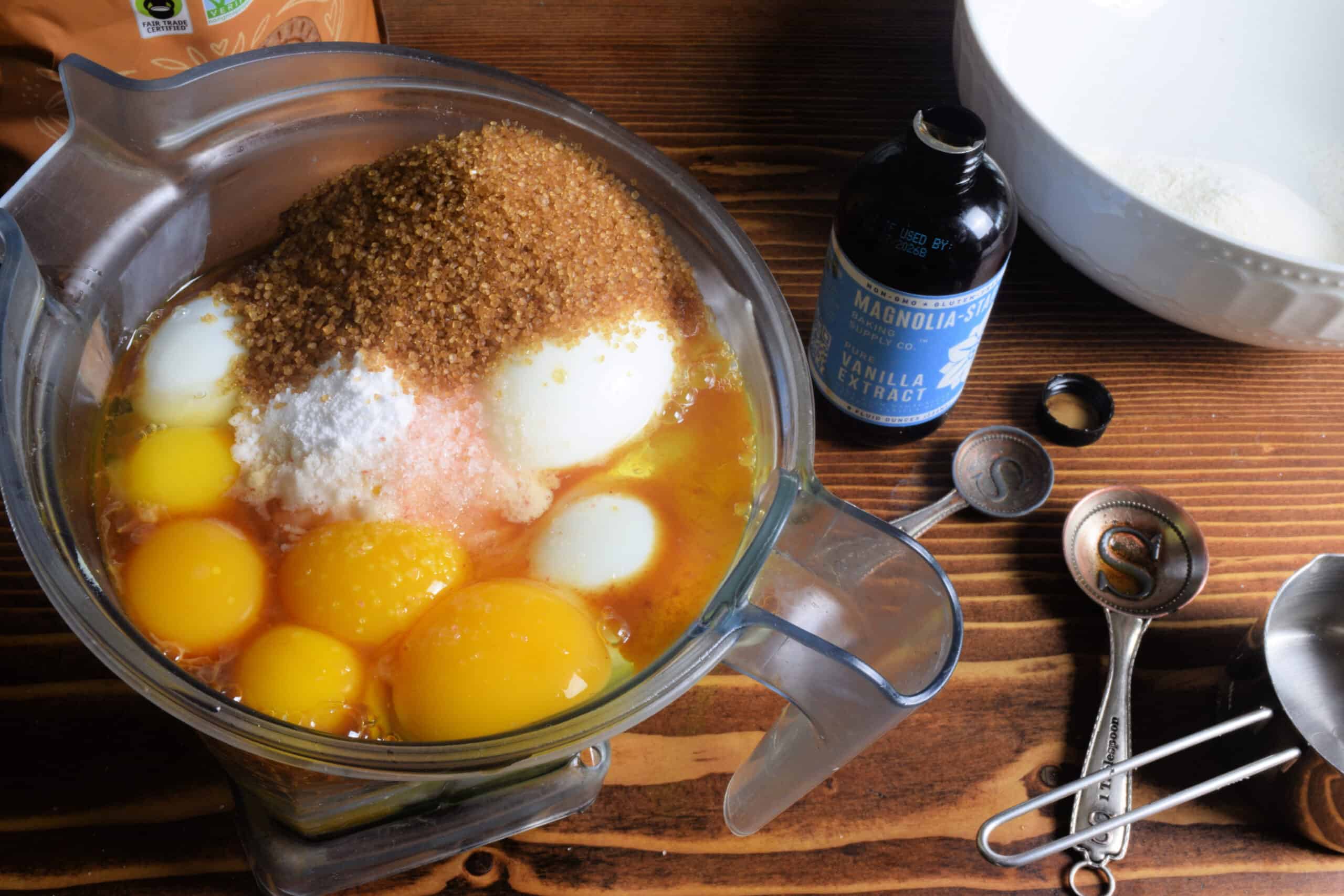 A blender on a wood countertop full of the ingredients for gluten free waffles that include: hard boiled eggs, raw eggs, sugar and coconut flour, next to vanilla