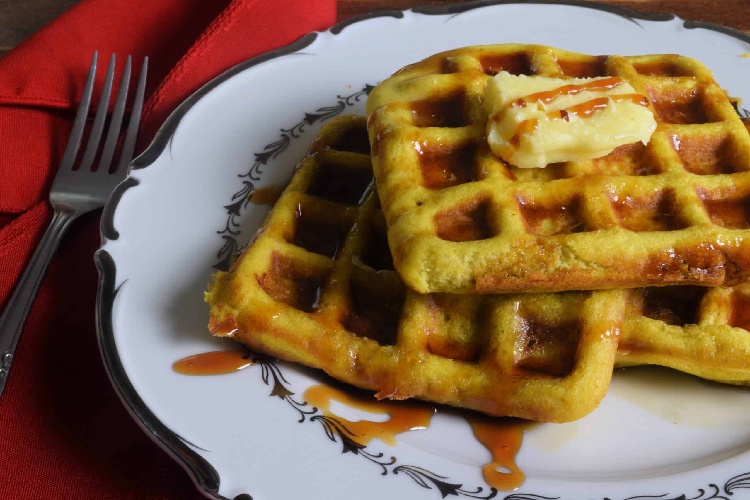 Gluten free waffles on a white plate covered in butter and maple syrup next to a red napkin.