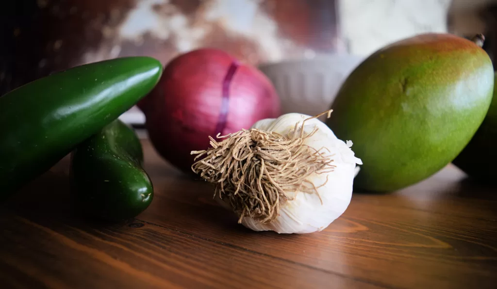 Ingredients for mango salsa: red onion, two jalapenos, garlic, two mango on a brown wood countertop.