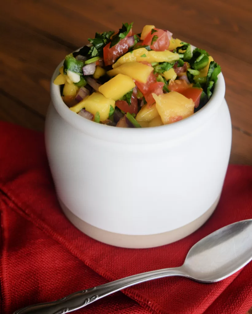 Easy mango salsa in a white serving jar with a red napkin beside it and a silver spoon on it, resting on a brown wood tabletop.