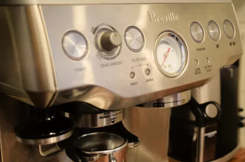 A clean Breville Barista Espresso Machine on a brown wood countertop next to a tower of coffee mugs, and a coffee bean grinder.