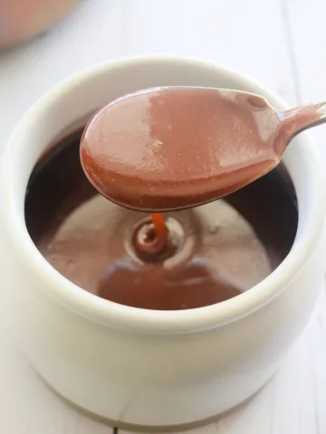 Spoon lifting glossy soft caramel sauce above a beige jar to show the thick, smooth texture.