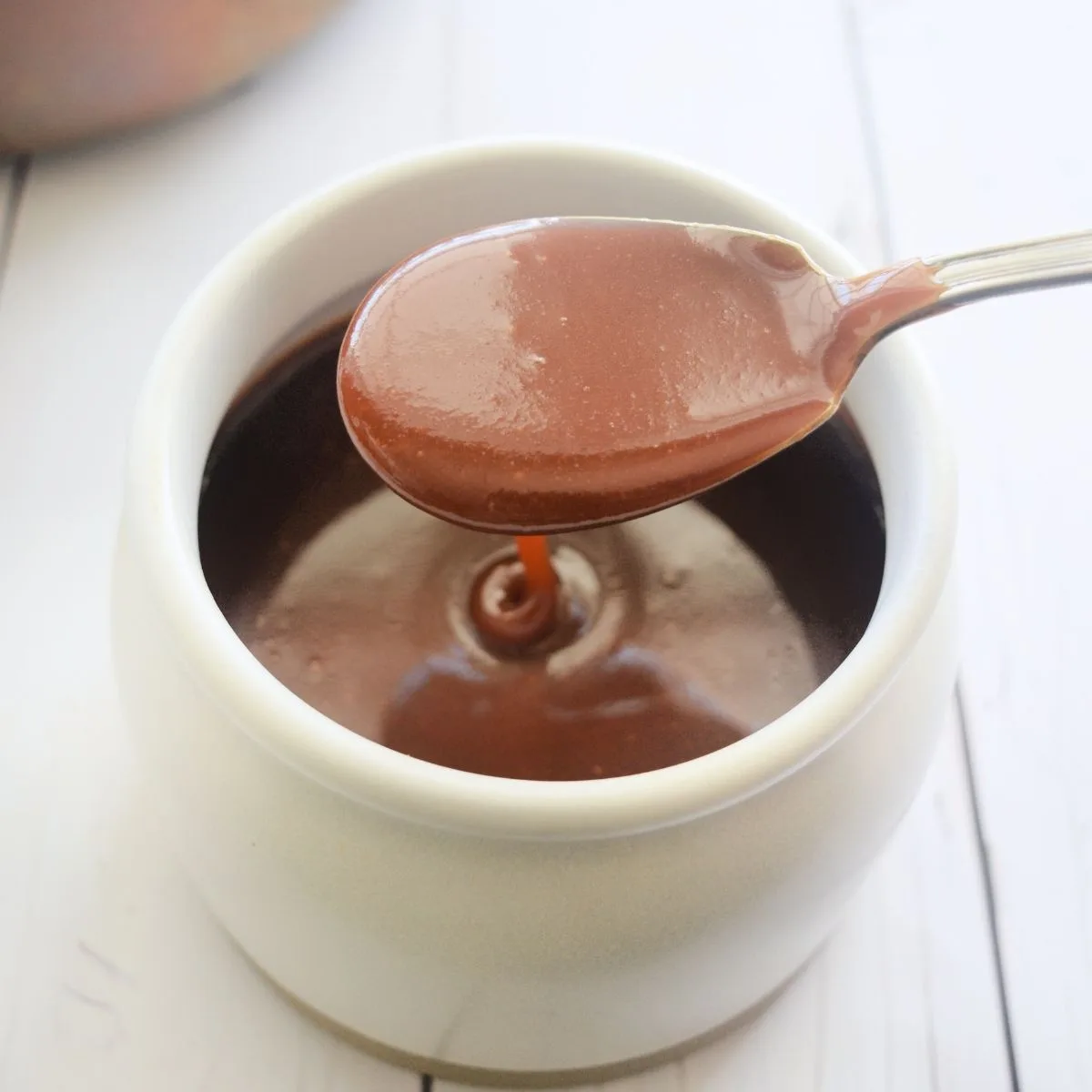 Spoon lifting glossy soft caramel sauce above a beige jar to show the thick, smooth texture.
