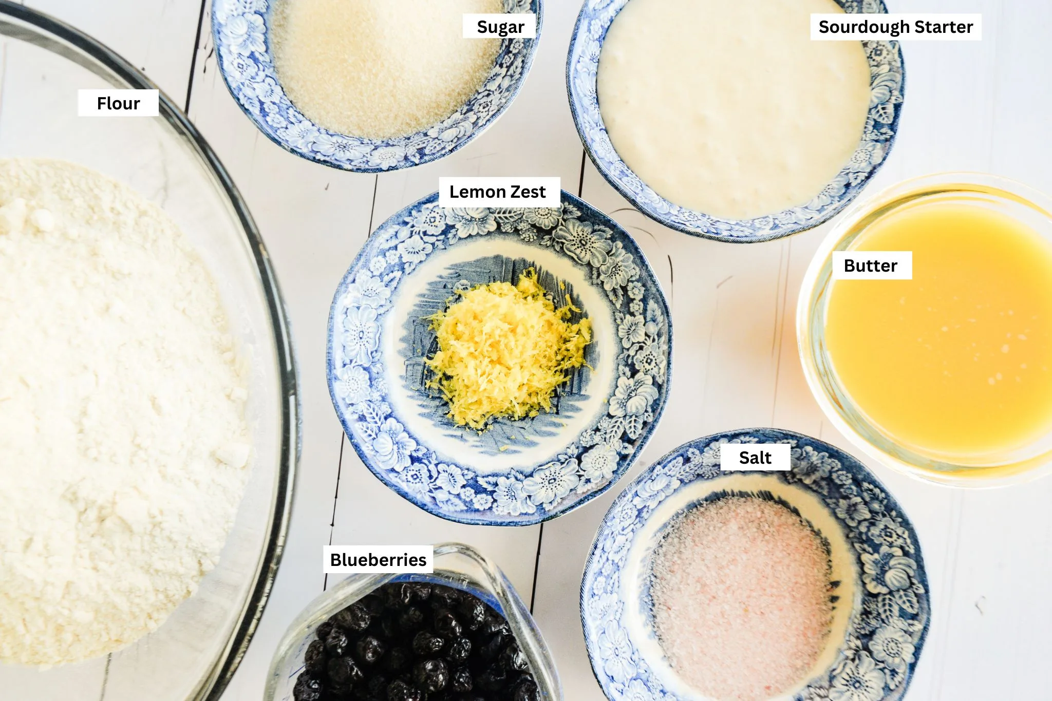 Overhead view of measured lemon blueberry sourdough bread ingredients, including flour, starter, sugar, lemon zest, blueberries, butter, and salt.