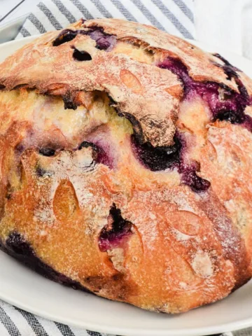 Close-up of a baked lemon blueberry sourdough loaf with decorative scoring, golden crust, and blueberries baked into the top.