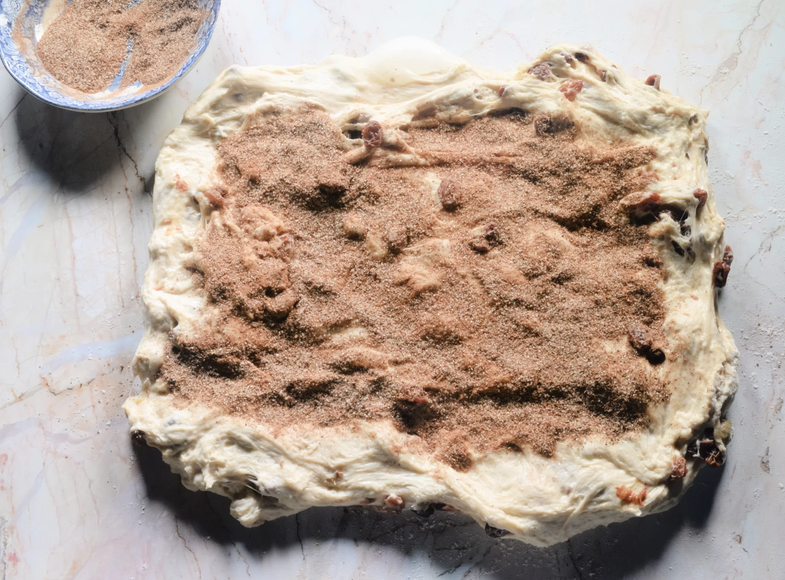 Cinnamon raisin sourdough dough stretched into a rough rectangle on a lightly floured marble surface, generously sprinkled with cinnamon sugar. A blue floral bowl with extra cinnamon sugar sits nearby, capturing a cozy old fashioned baking step in the recipe.
