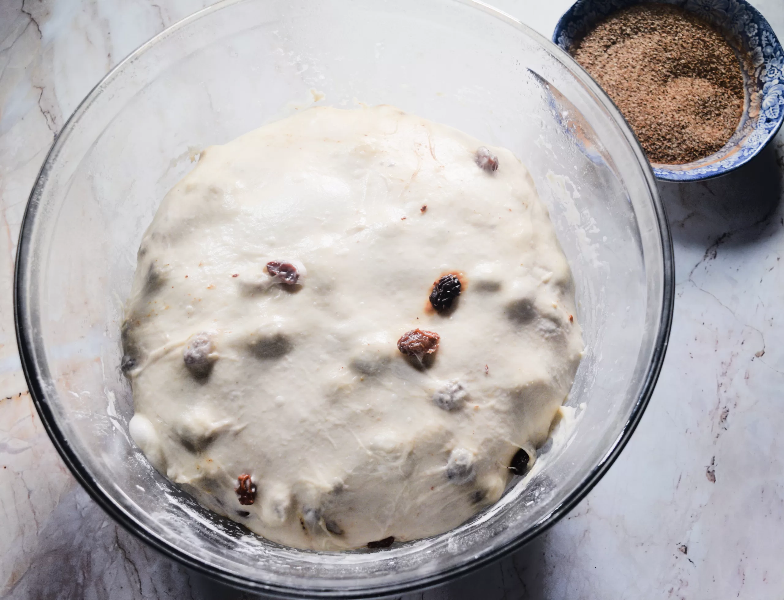 Overhead view of cinnamon raisin sourdough dough after rising in a clear glass bowl, with raisins peeking through the smooth surface. A small blue bowl of cinnamon sugar sits nearby on a marble countertop, creating a warm old fashioned baking scene.