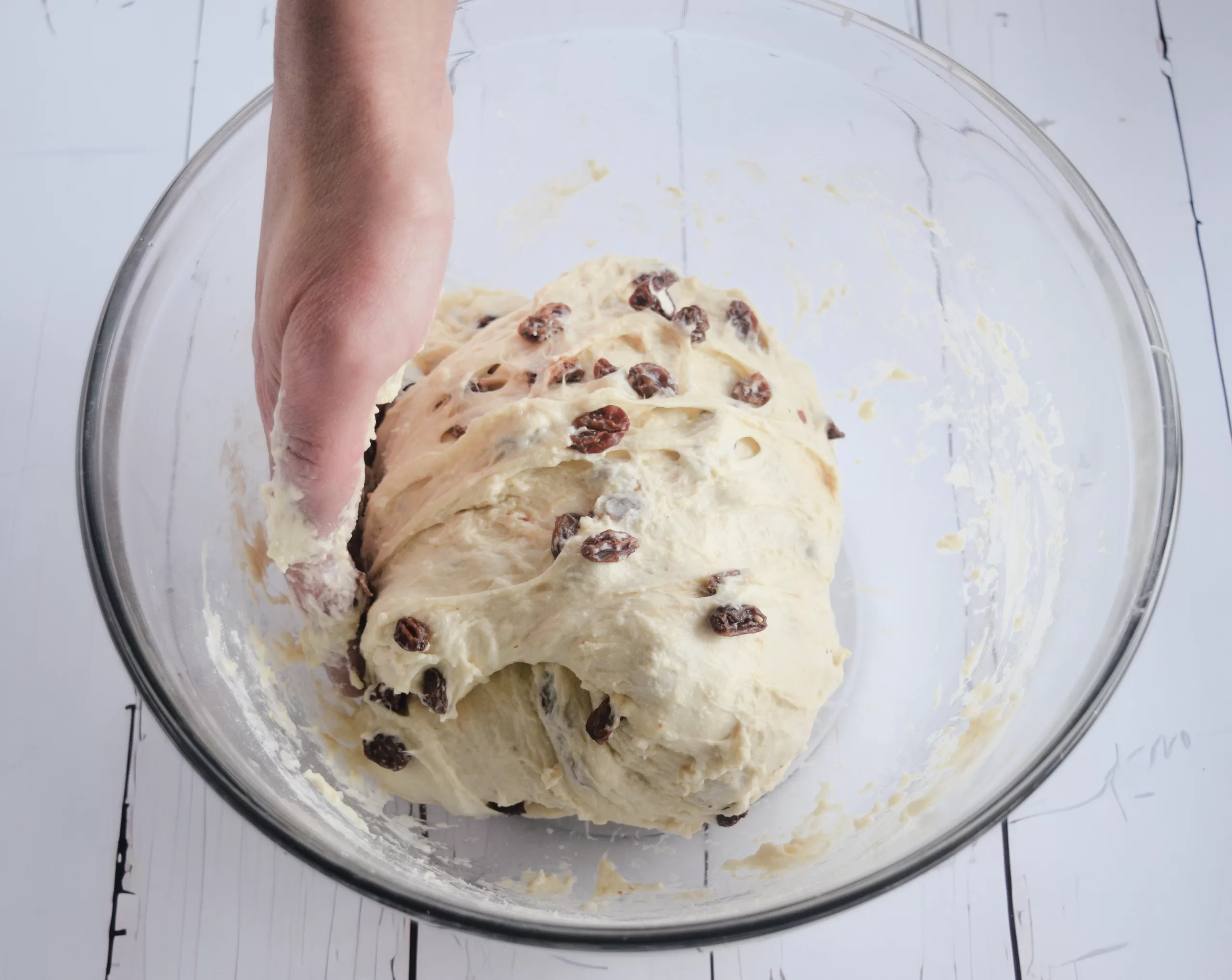 Hand folding a soft ball of cinnamon raisin sourdough dough inside a clear glass bowl. Raisins are visible throughout the dough, shown mid-fold on a white wooden surface for a cozy old fashioned baking process.