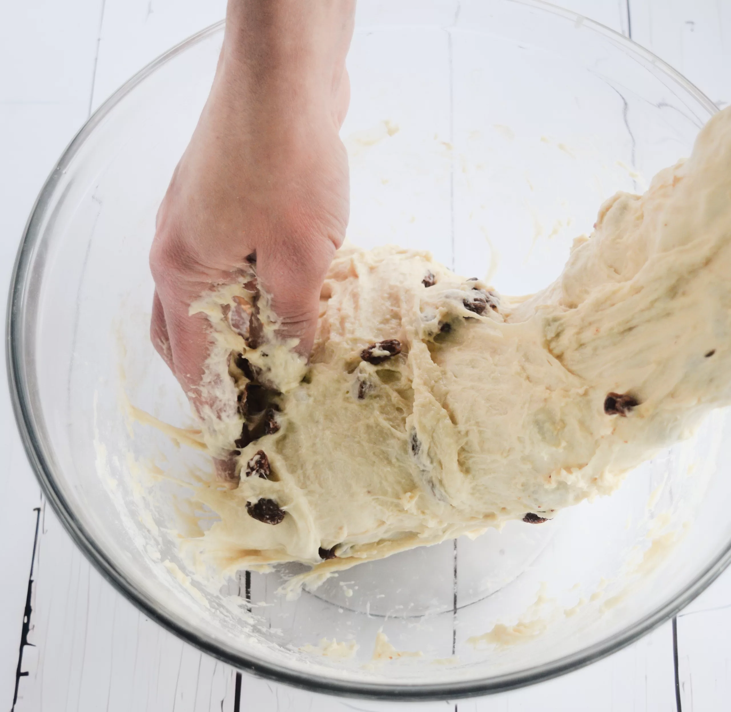 “Hand lifting and stretching cinnamon raisin sourdough dough inside a clear glass bowl. The dough is soft and elastic with raisins distributed throughout, shown during a fold on a white wooden surface in a simple old fashioned kitchen setting.