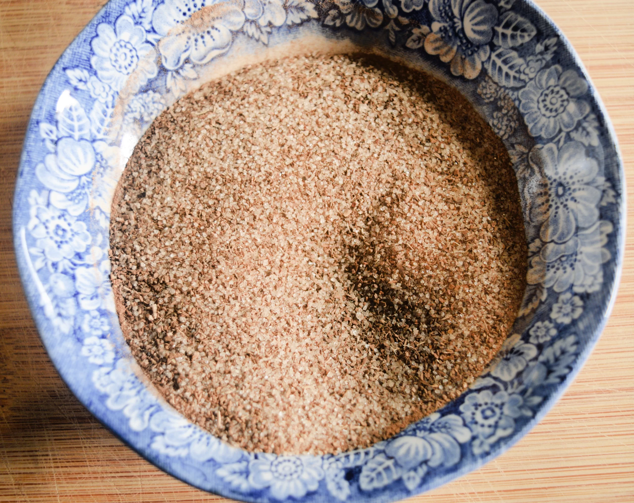 Overhead view of a blue and white floral bowl filled with a cinnamon sugar mixture. The fine, sandy blend sits on a wooden surface, ready to be used in a homemade cinnamon raisin sourdough bread recipe.