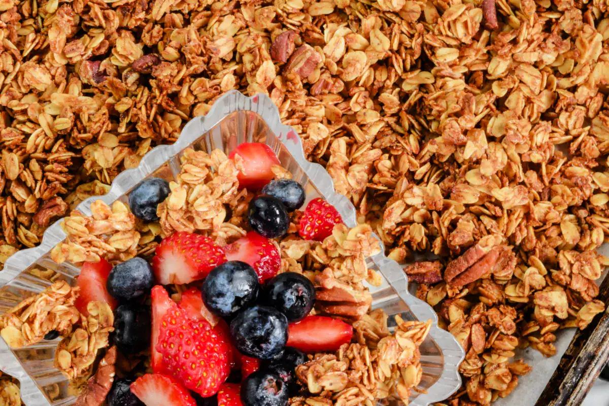 Homemade granola with fresh strawberries and blueberries served in a clear dish on a baking sheet lined with golden, crunchy oat clusters and pecans—perfect healthy breakfast or snack option.