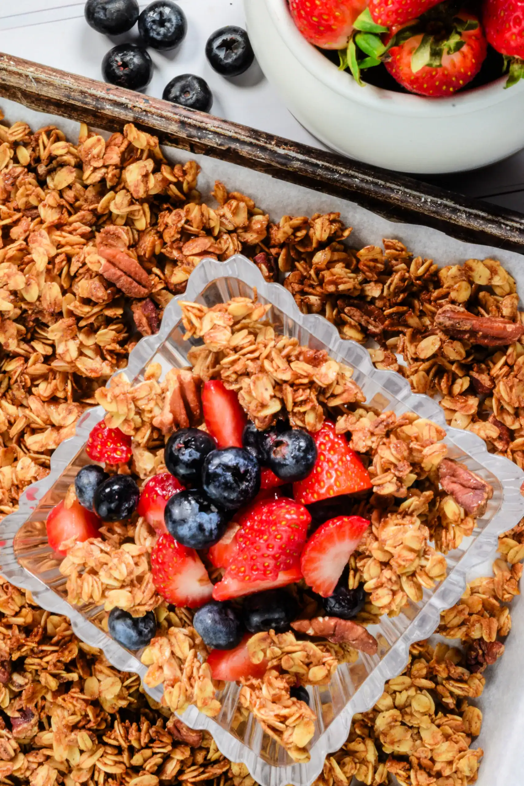 Homemade granola with fresh strawberries and blueberries served in a clear dish on a baking sheet lined with golden, crunchy oat clusters and pecans—perfect healthy breakfast or snack option.