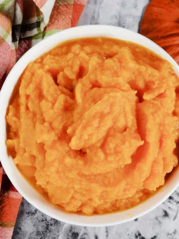 Homemade pumpkin purée in a white bowl with a plaid towel and whole pumpkin nearby.