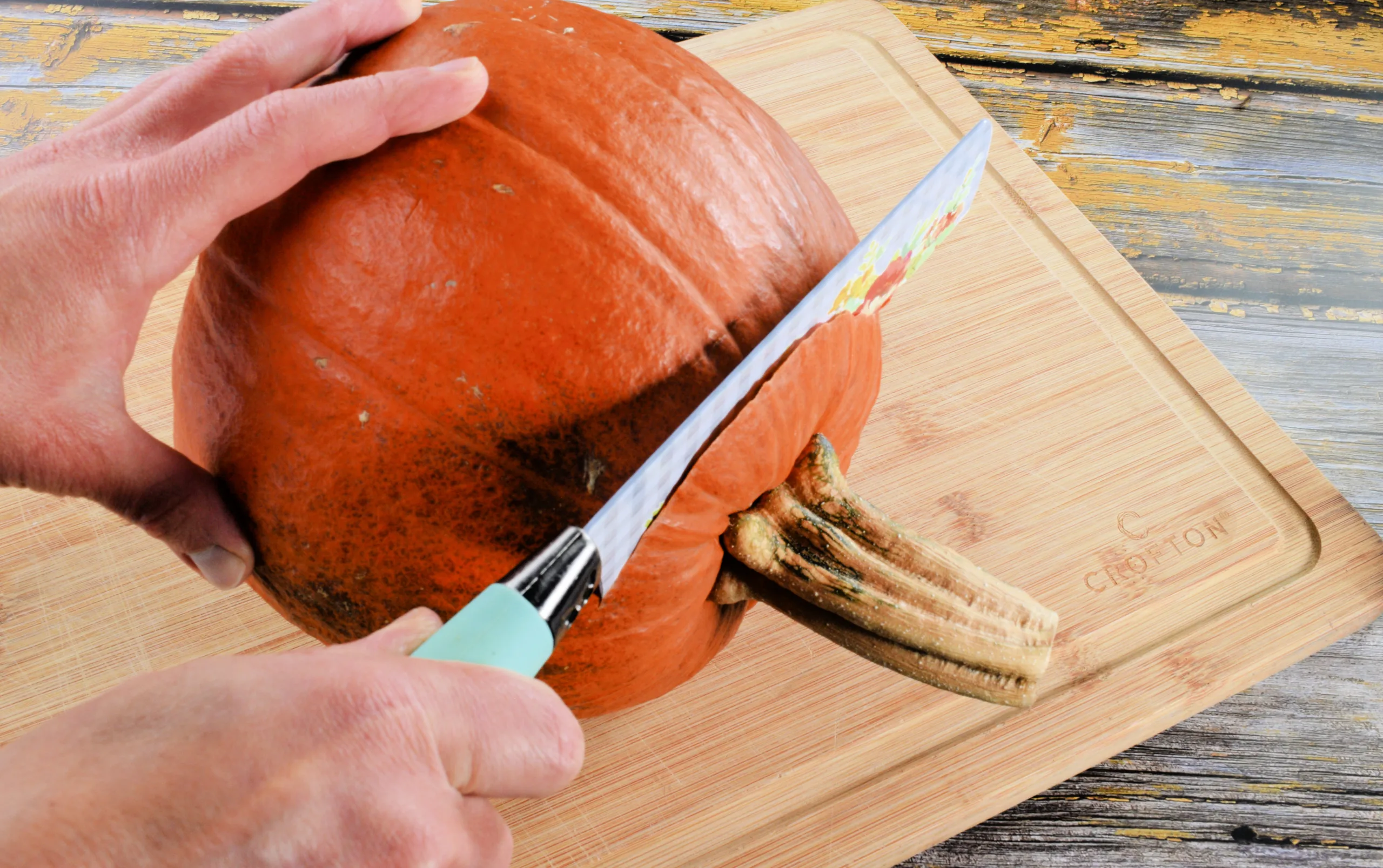 Whole pumpkin being cut open with a sharp knife on a wooden cutting board.