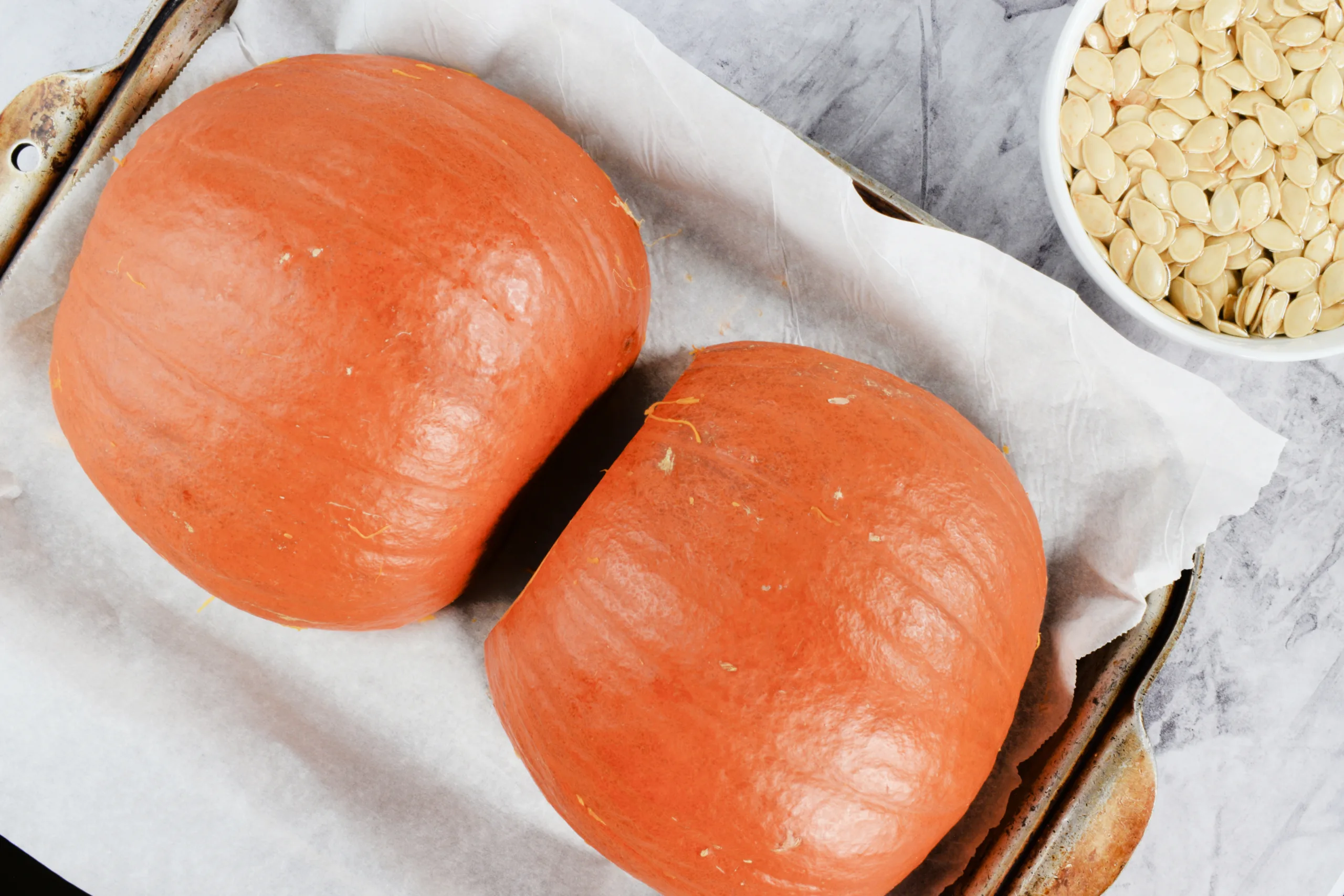 Pumpkin halves cut side down on a parchment-lined baking sheet with cleaned pumpkin seeds nearby.
