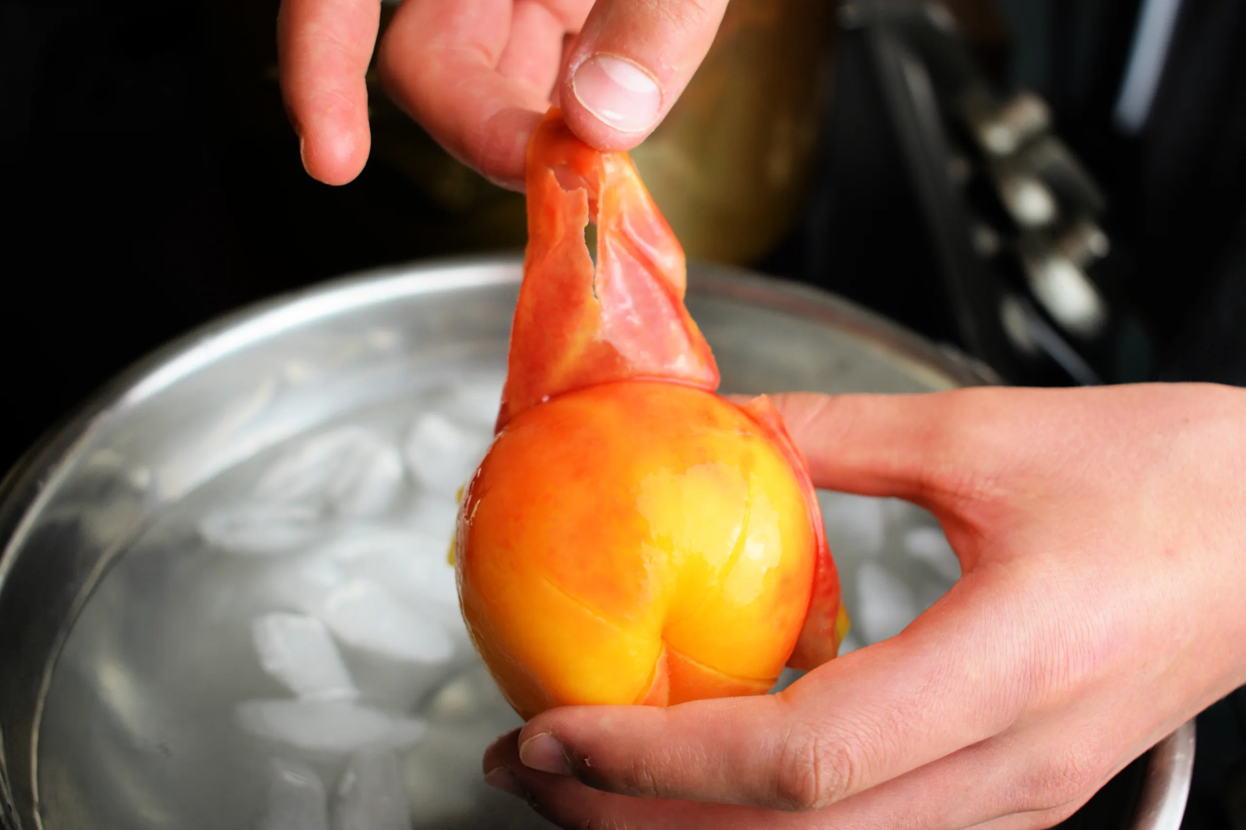 Skin being peeled from a blanched peach over a bowl of ice water.