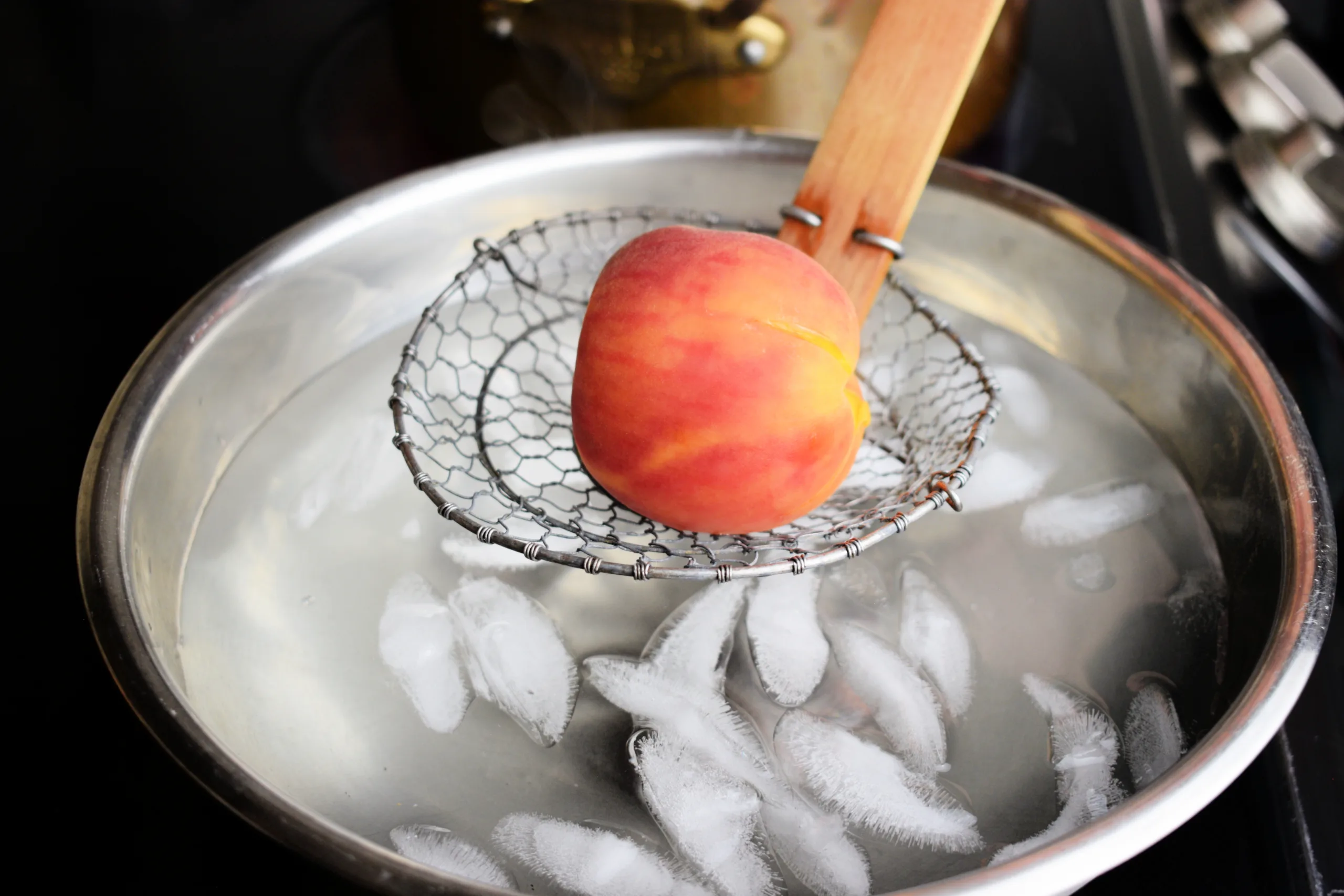 Blanched peach resting on a wire strainer over an ice bath before peeling.