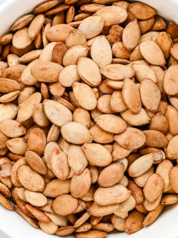 Roasted pumpkin seeds in a white bowl with golden brown edges.