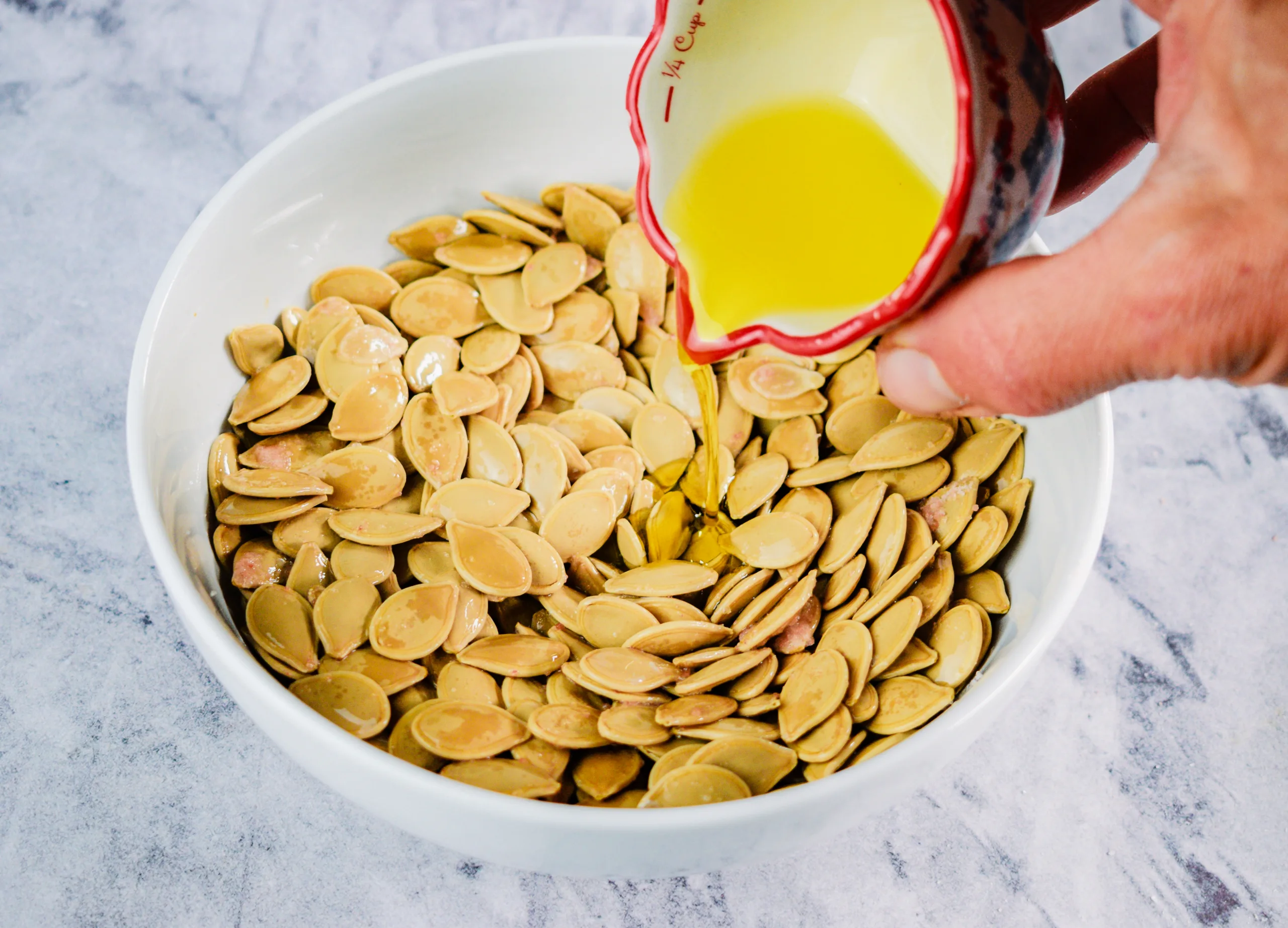 Olive oil being poured over clean pumpkin seeds in a white bowl.