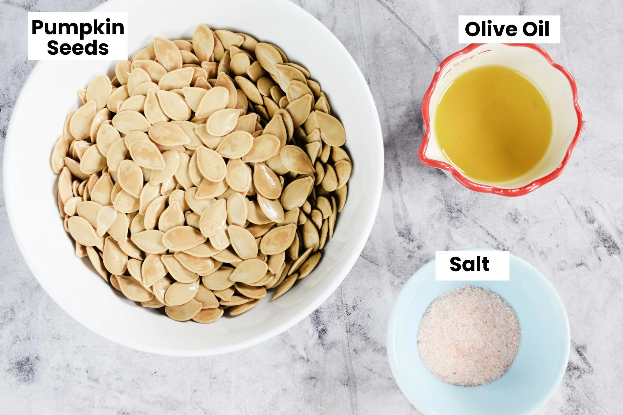 Pumpkin seeds, olive oil, and salt arranged on a gray surface with ingredient labels for roasted pumpkin seeds.