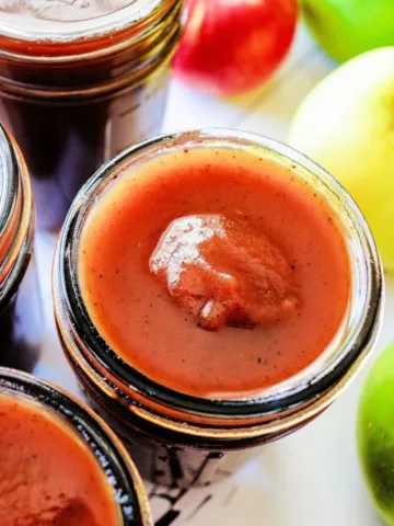 Thick homemade apple butter in a glass jar with fresh red and green apples behind it.