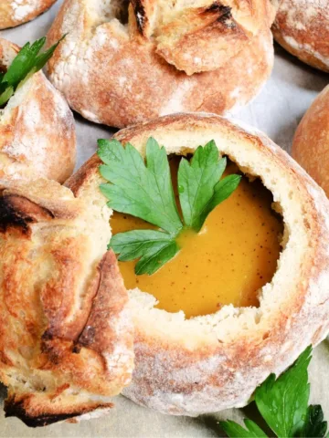 Crusty homemade sourdough bread bowl filled with creamy orange soup and topped with a fresh parsley leaf, surrounded by more sourdough bread bowls on parchment paper.