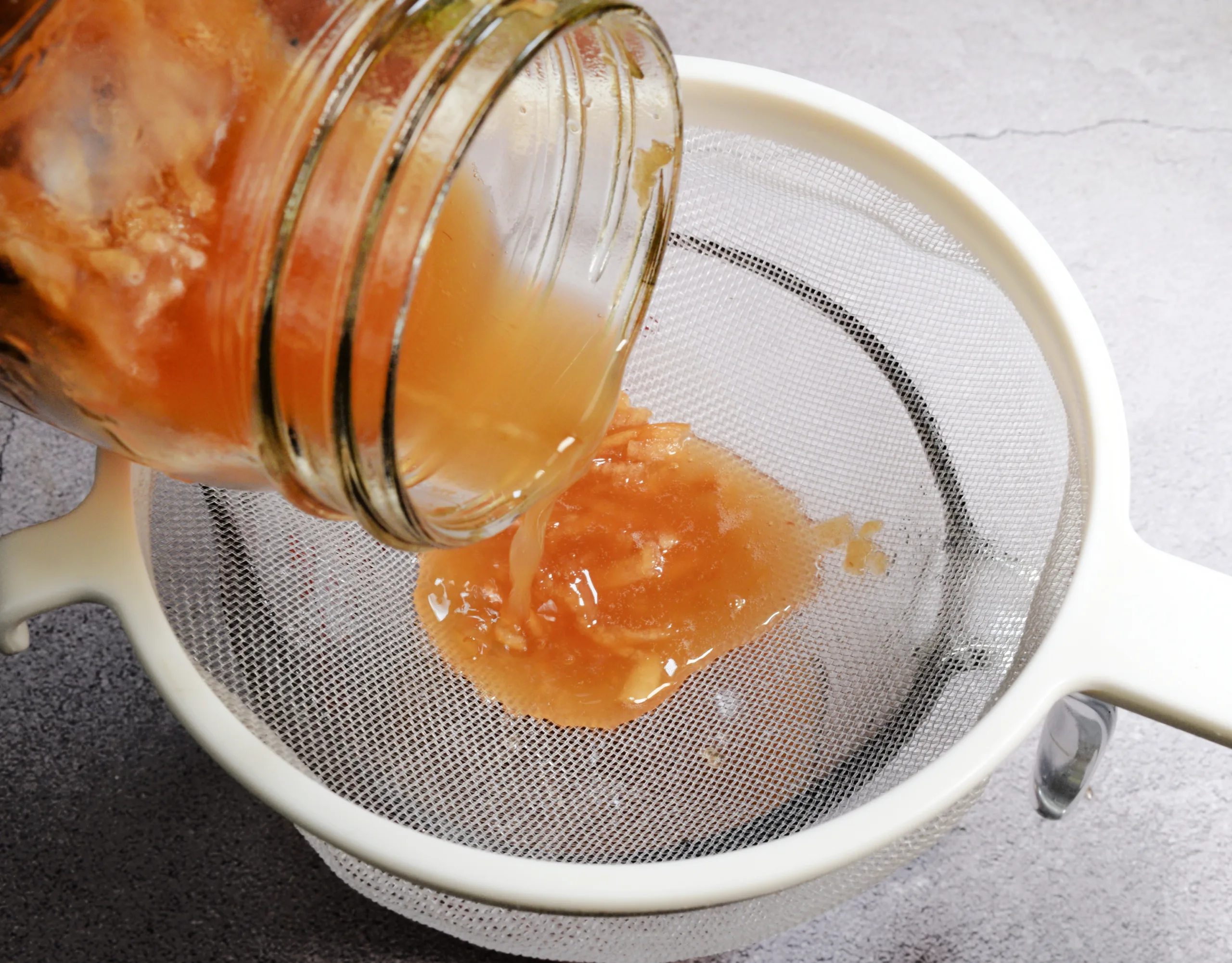 Apple cinnamon shrub being poured from a mason jar through a fine mesh strainer.