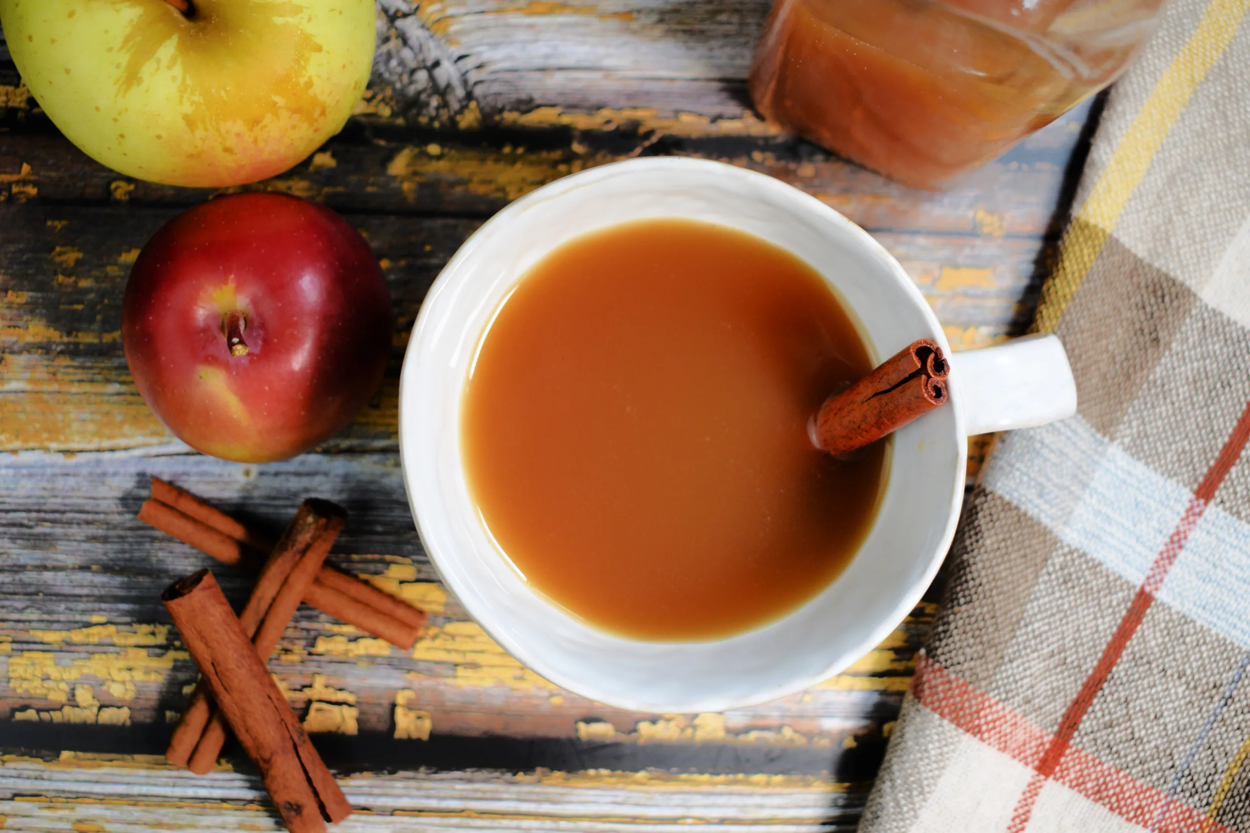 Apple cinnamon shrub in a white mug with a cinnamon stick, fresh apples, a jar, and a plaid towel.