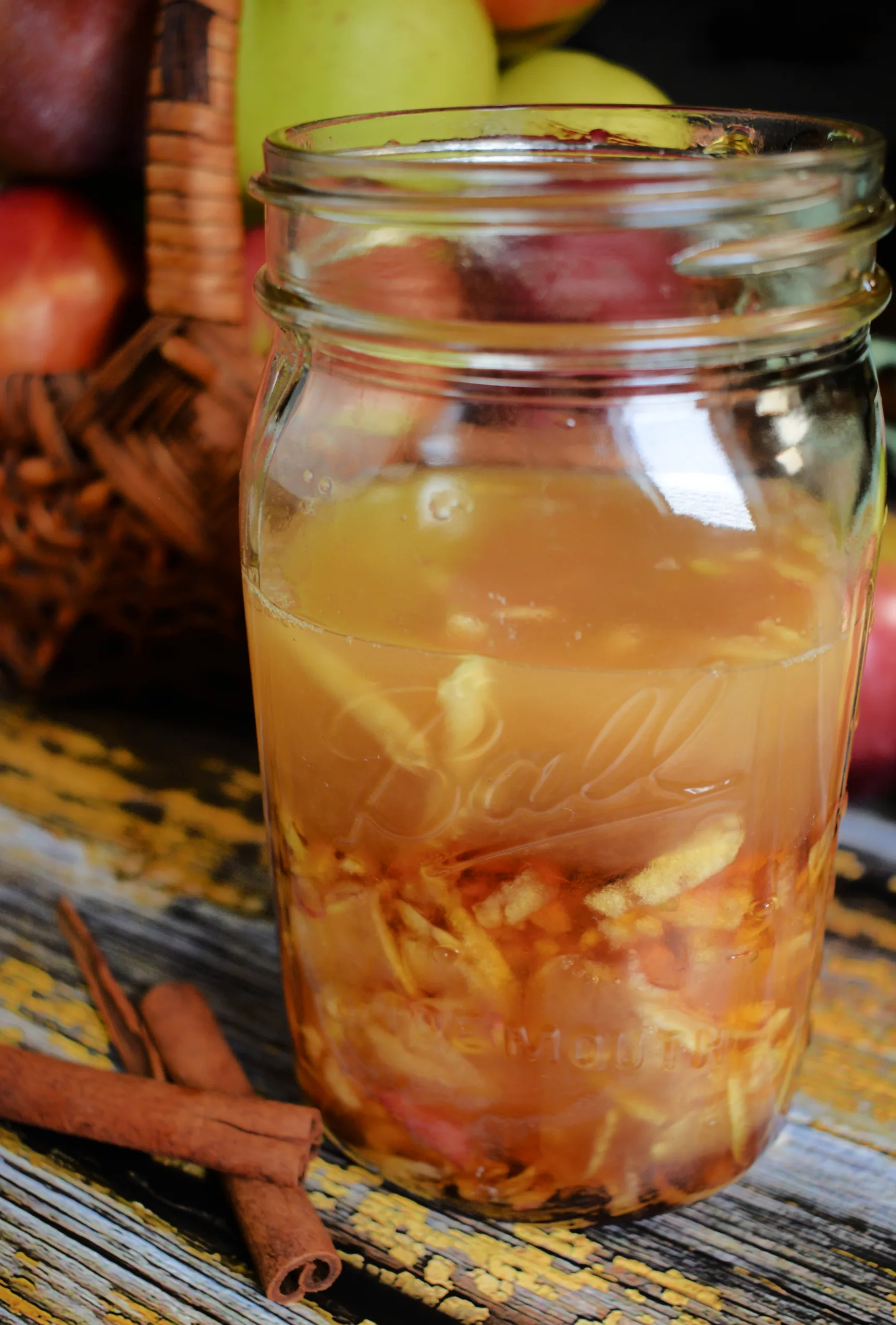 A glass bottle filled with homemade apple cinnamon shrub, with apples, cinnamon sticks, and a apples behind it.