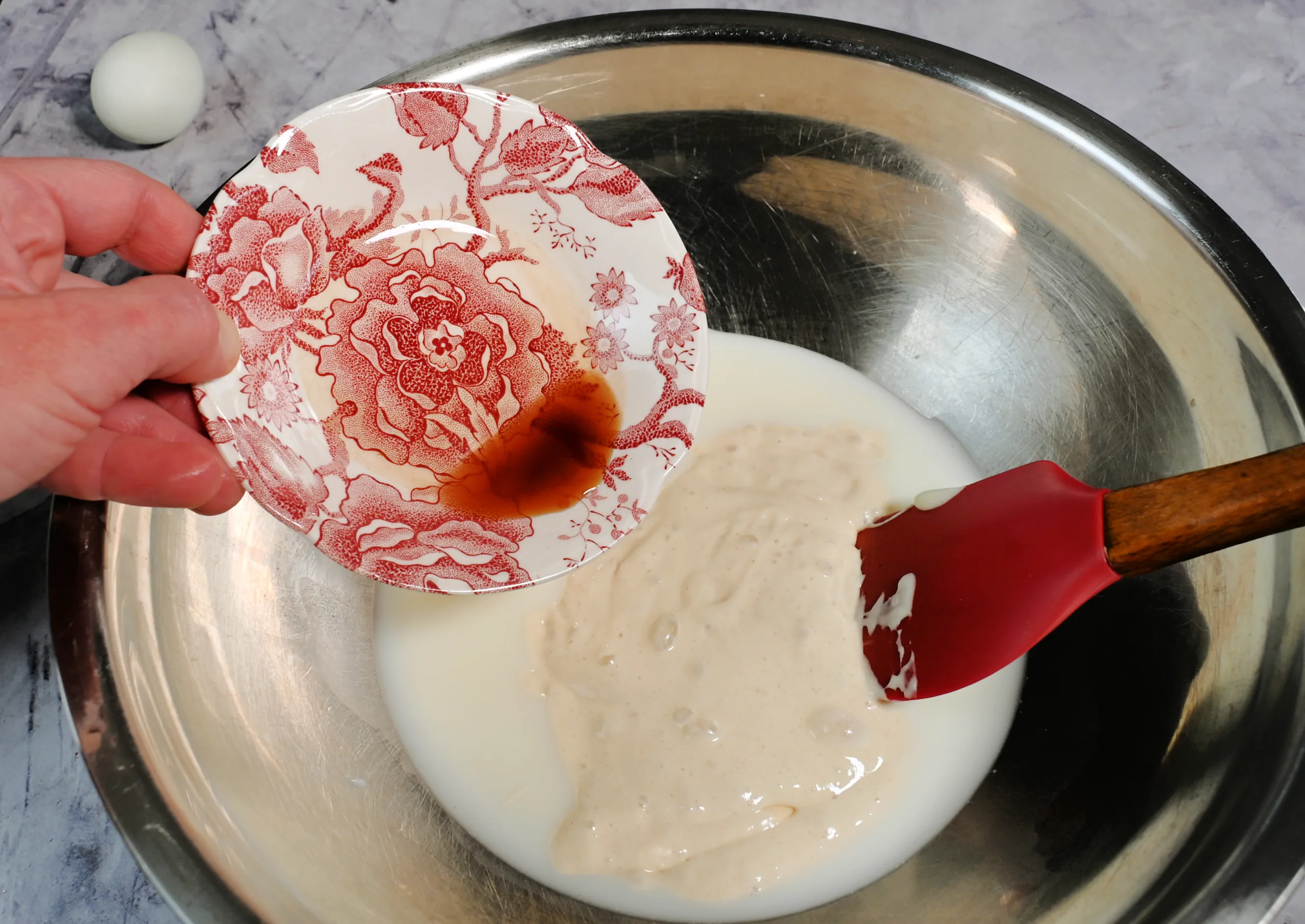Hand pouring vanilla extract from a small red floral dish into a mixing bowl filled with sourdough discard, milk, and a red spatula. Overhead view on a marble countertop, capturing the first steps of making fluffy sourdough discard pancakes in a cozy, old fashioned kitchen setting.
