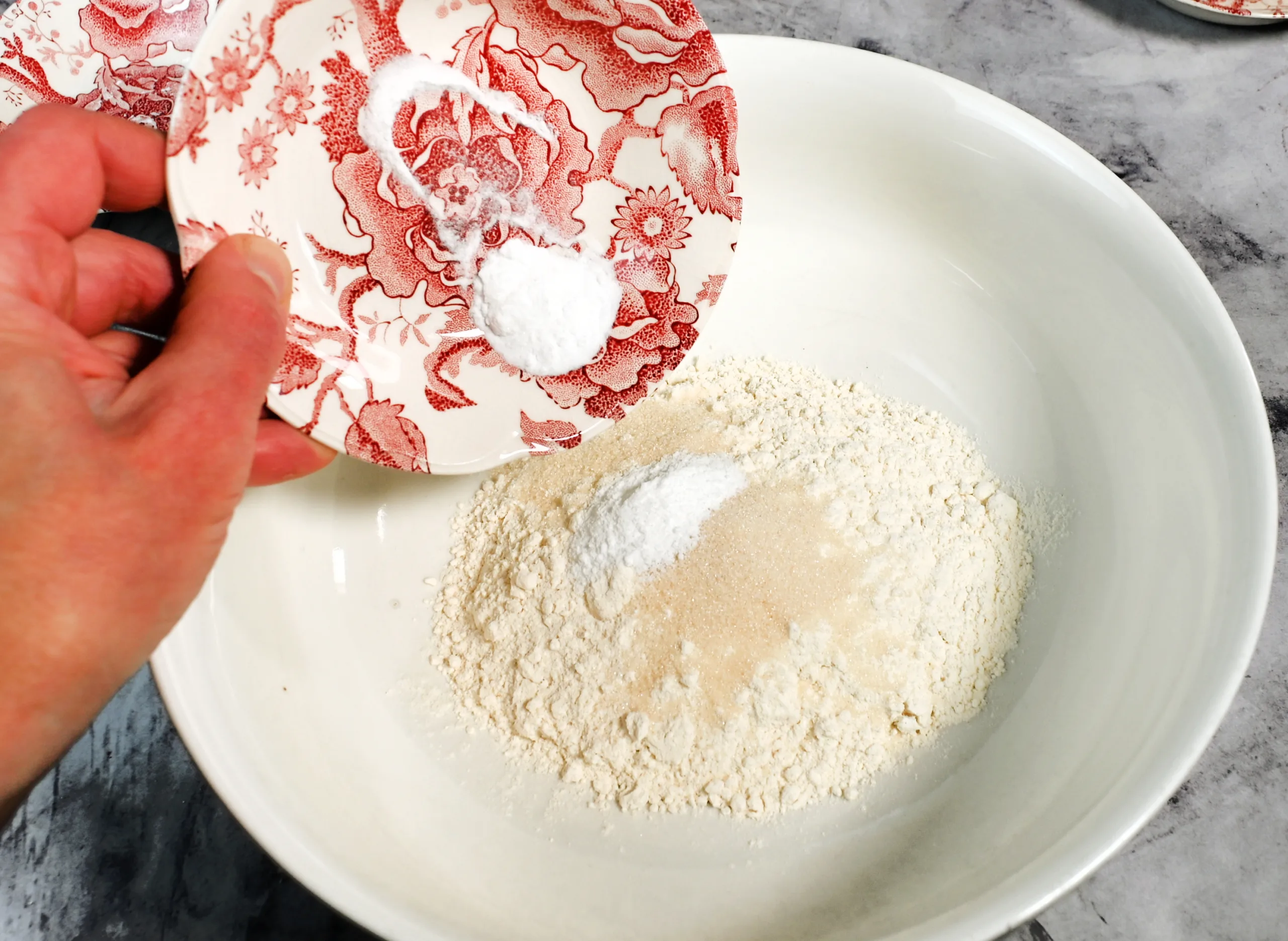 Hand holding a small red floral dish and adding baking soda and baking powder to a bowl of flour on a marble countertop. Overhead view of the dry ingredients for fluffy sourdough discard pancakes being combined in a simple, old fashioned kitchen setting.