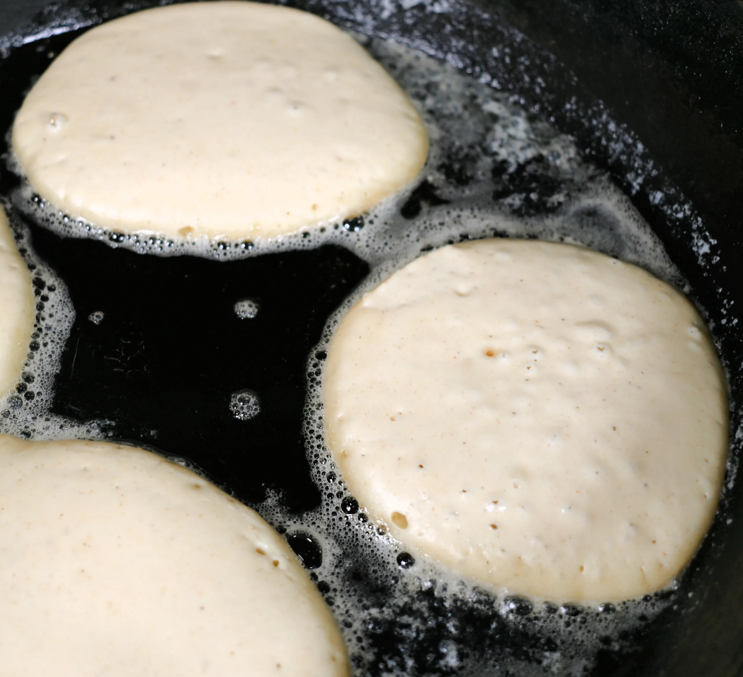 Fluffy sourdough discard pancake batter cooking in a hot buttered skillet, with small bubbles forming on the surface as the edges begin to set. Close up view capturing the classic look of pancakes just before flipping in a cozy, old fashioned kitchen setting.
