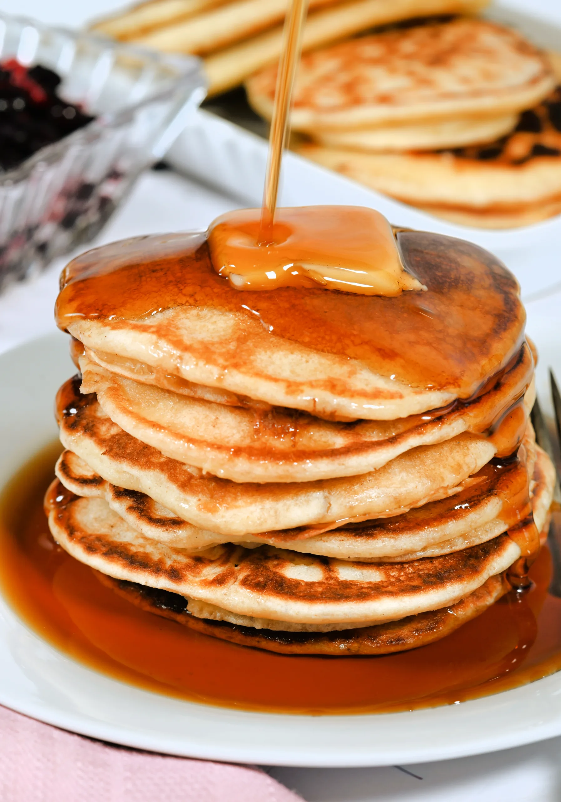 Tall stack of fluffy sourdough discard pancakes topped with a pat of butter as warm maple syrup is poured over the top. Golden edges and soft centers are visible, with extra pancakes and a bowl of berries in the background for a cozy old fashioned breakfast scene.