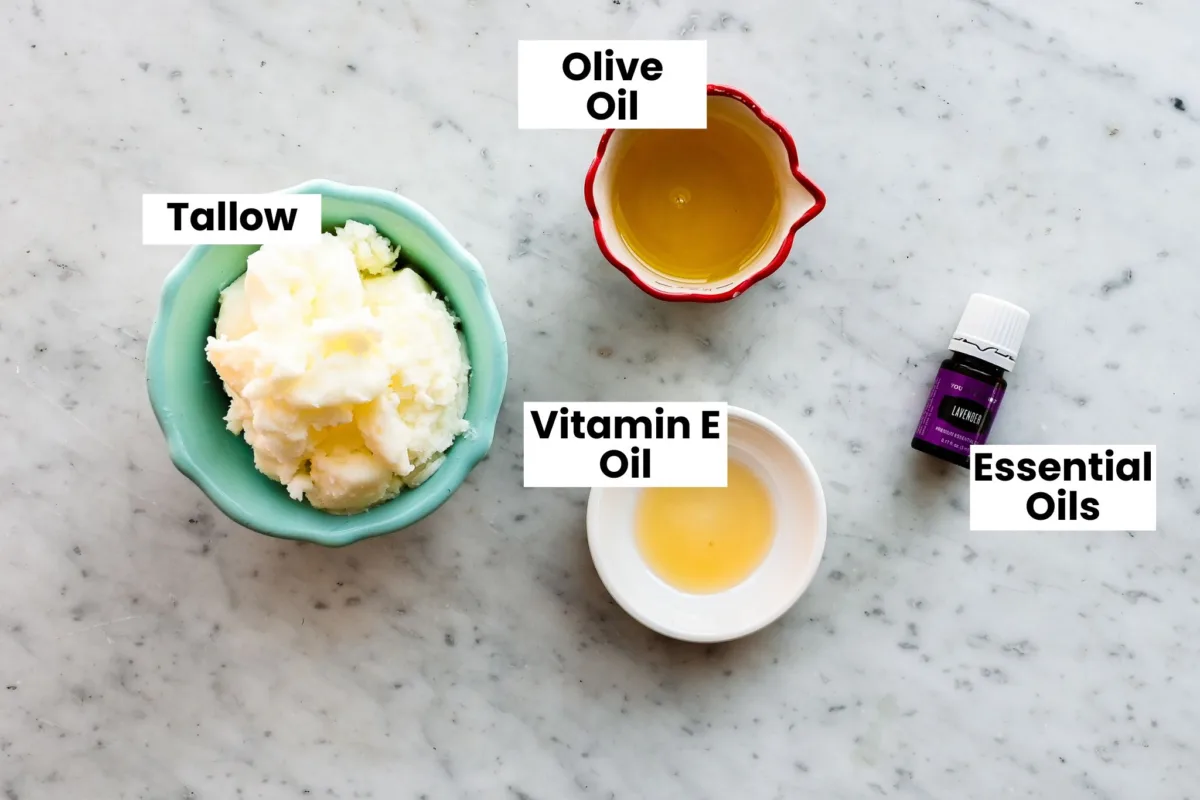 Grass-fed tallow, olive oil, vitamin E oil, and essential oils arranged on a marble surface for making a homemade whipped tallow balm using a simple DIY recipe.