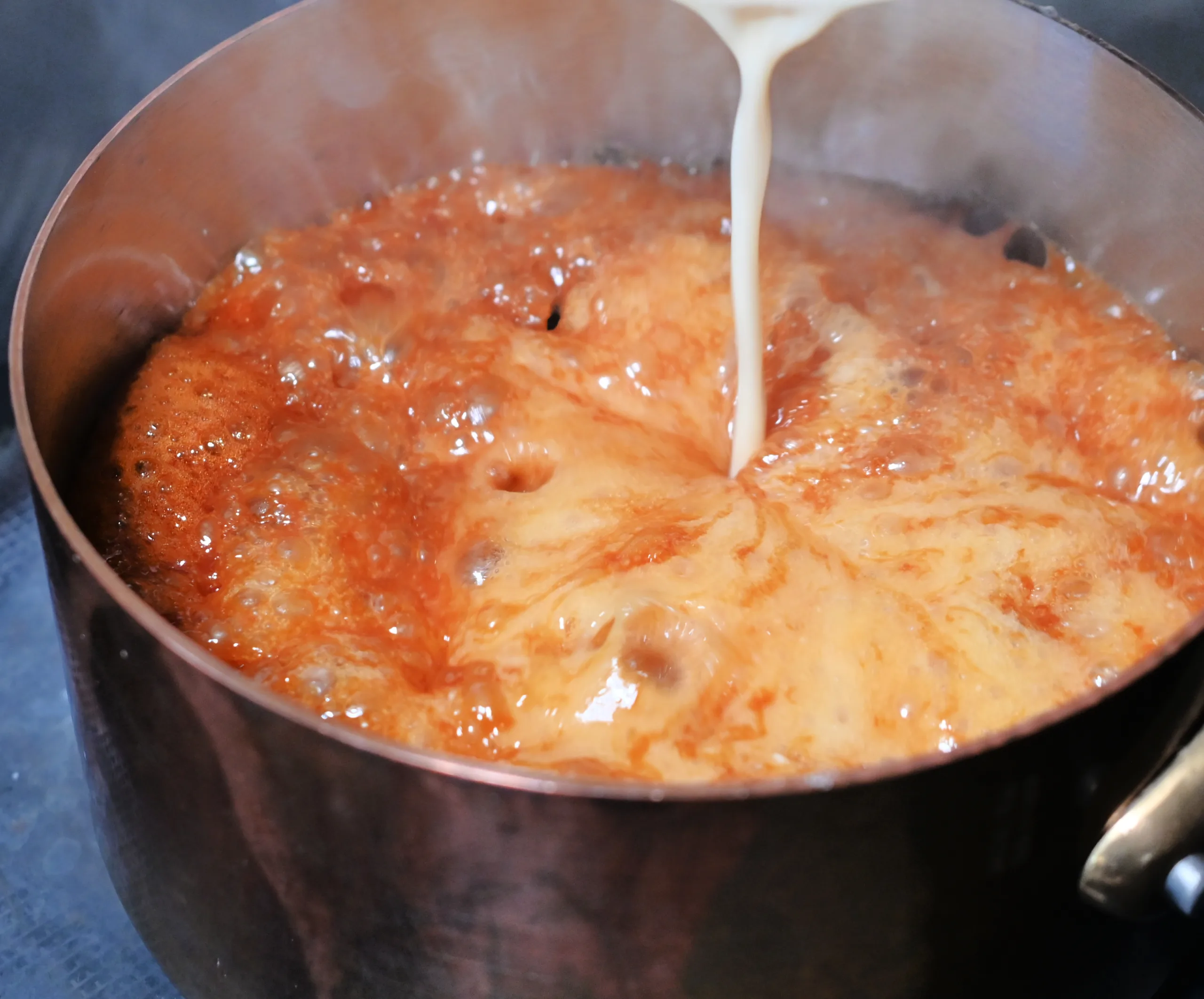 Warm cream being poured into caramelized sugar for homemade caramel candy.