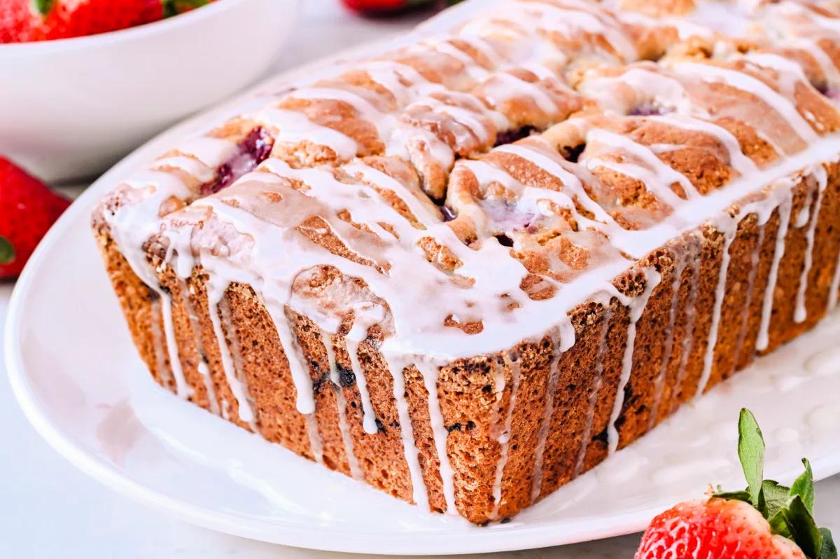 A close-up of a freshly baked loaf of strawberry sourdough quick bread with a golden-brown crust, drizzled with a white glaze that cascades down the sides. The loaf has a slightly cracked top, revealing bits of strawberries inside. It sits on a white plate with a few fresh strawberries scattered around, adding a pop of red color in the background.