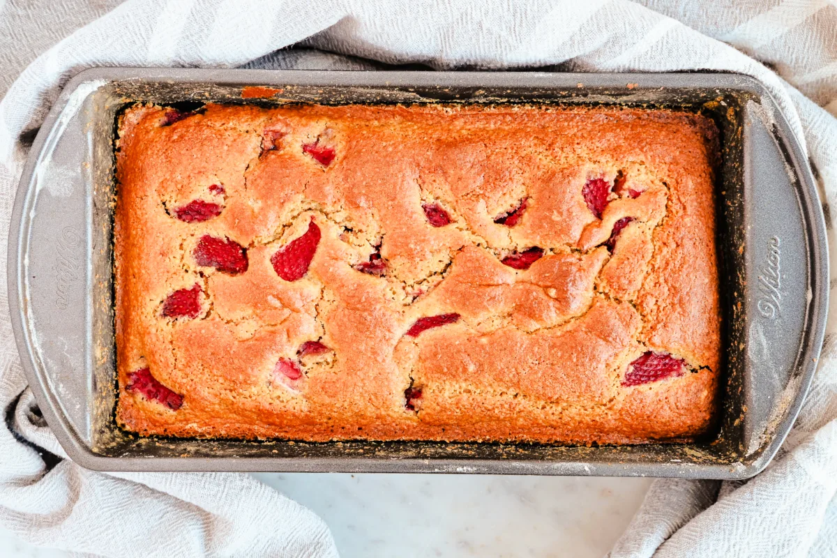 A freshly baked loaf of strawberry sourdough quick bread in a dark metal baking pan. The golden-brown crust has a slightly cracked surface, revealing chunks of bright red strawberries baked into the bread. The pan sits on a marble countertop with a light-colored kitchen towel draped around it.
