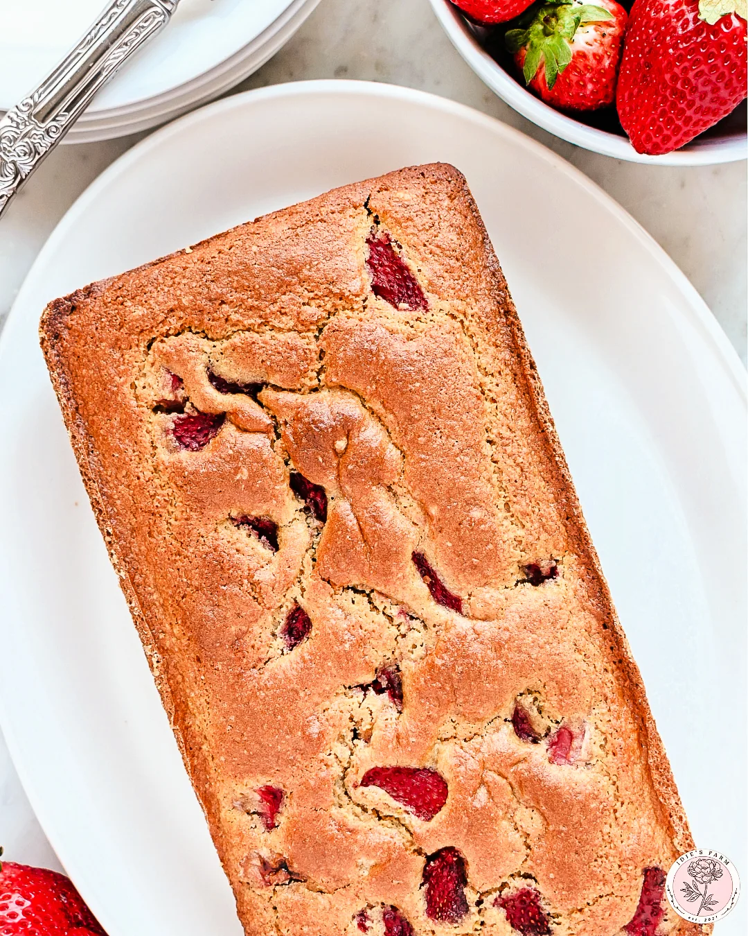 A freshly baked loaf of strawberry sourdough quick bread in a dark metal baking pan. The golden-brown crust has a slightly cracked surface, revealing chunks of bright red strawberries baked into the bread. The pan sits on a marble countertop with a light-colored kitchen towel draped around it.