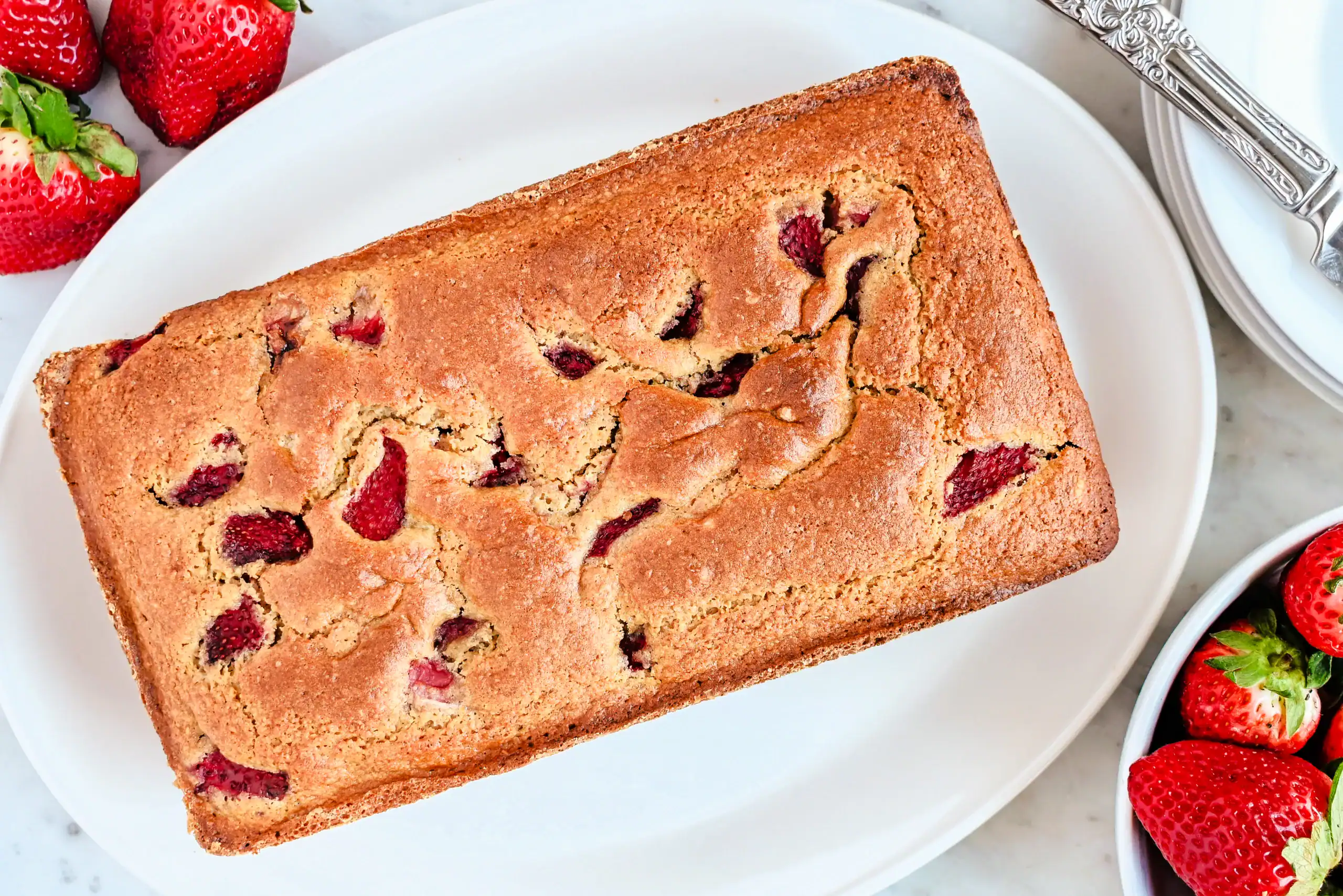 A freshly baked loaf of strawberry sourdough quick bread in a dark metal baking pan. The golden-brown crust has a slightly cracked surface, revealing chunks of bright red strawberries baked into the bread. The pan sits on a marble countertop with a light-colored kitchen towel draped around it.
