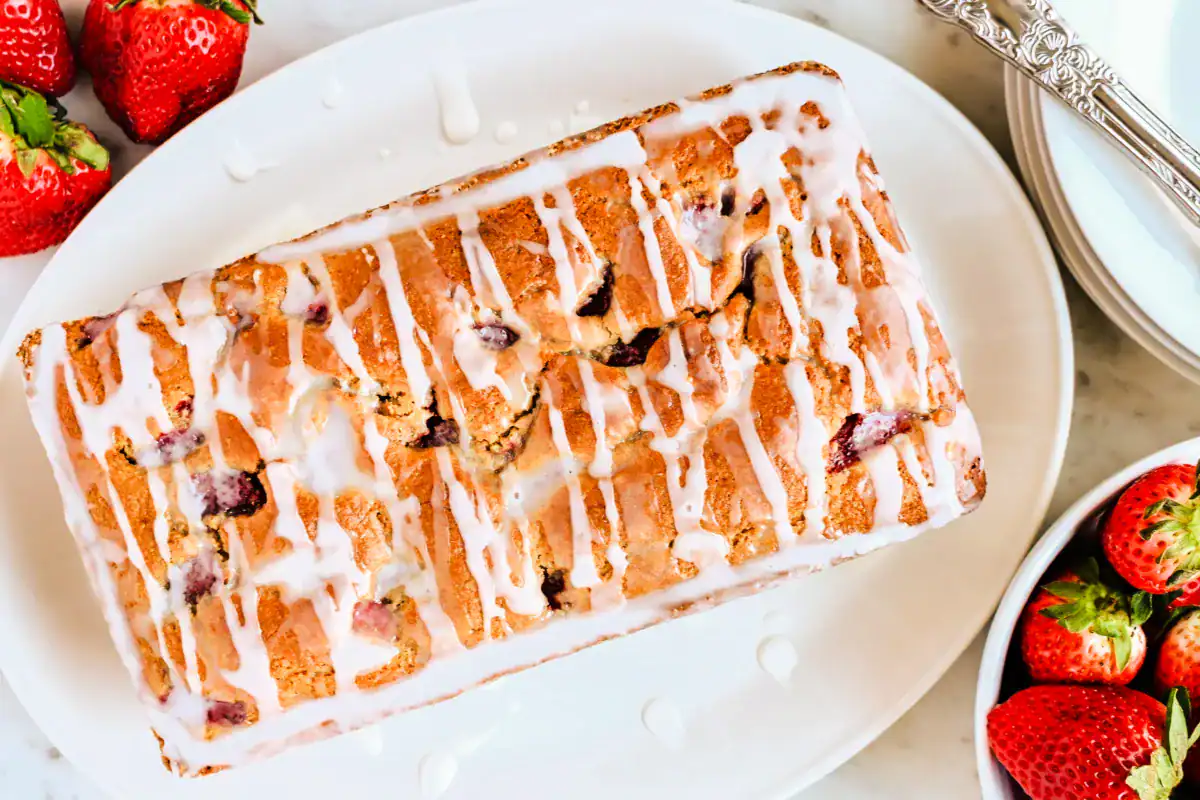 A close-up of a freshly baked loaf of strawberry sourdough quick bread with a golden-brown crust, drizzled with a white glaze that cascades down the sides. The loaf has a slightly cracked top, revealing bits of strawberries inside. It sits on a white plate with a few fresh strawberries scattered around, adding a pop of red color in the background.